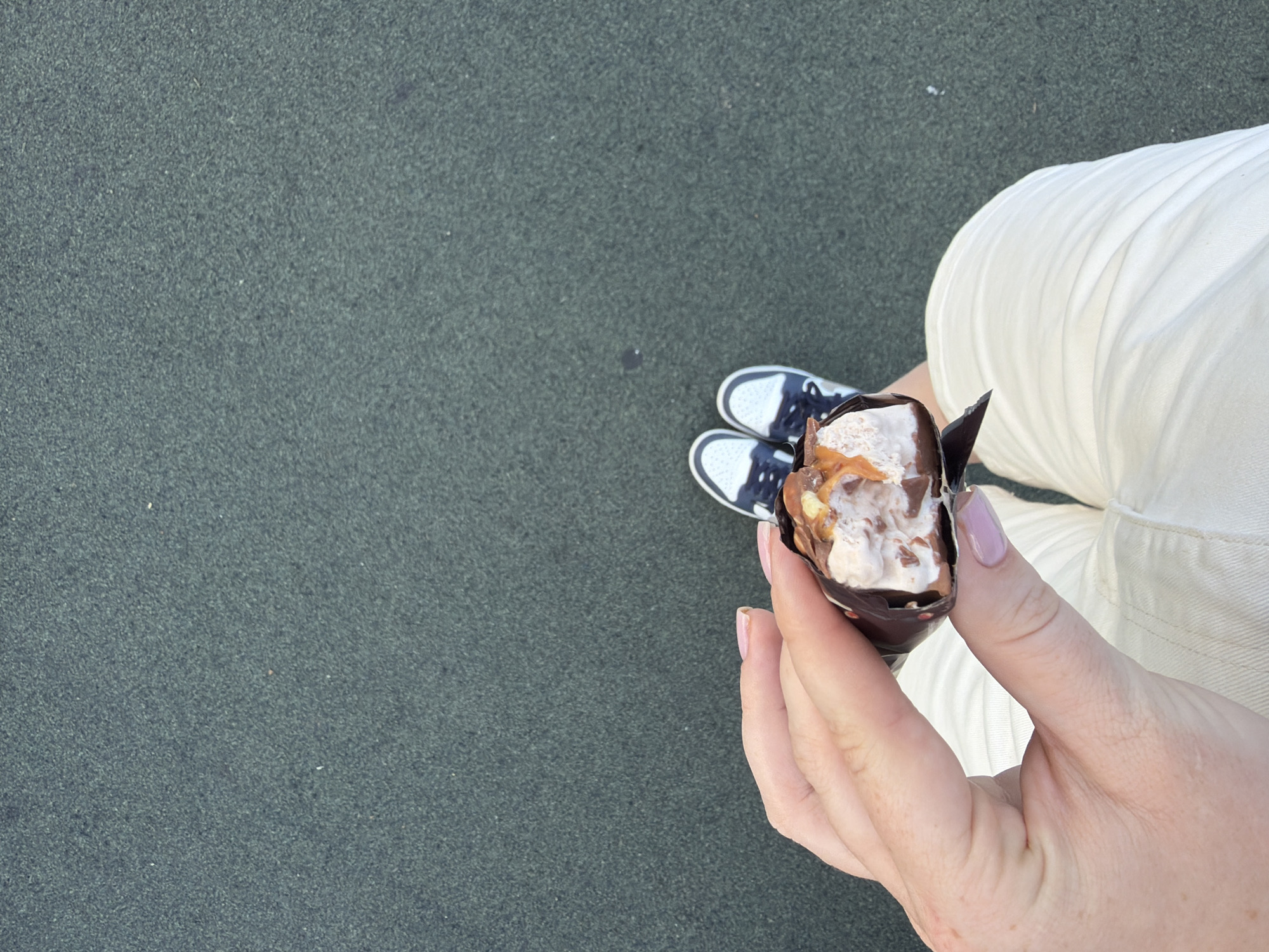 In this image, a person is holding a partially eaten ice cream bar. The ice cream has a chocolate coating and appears to have nougat and caramel filling. The person's hand, with neatly manicured nails, is visible in the foreground. They are wearing white shorts and athletic shoes. The background consists of a dark, textured surface, likely asphalt or a similar material common in urban outdoor areas. The image is taken from a high-angle perspective, looking down on the subject. The scene is suggestive of a casual outdoor setting, possibly a park or a street in Kazan, Russia, where such snacks are commonly consumed.