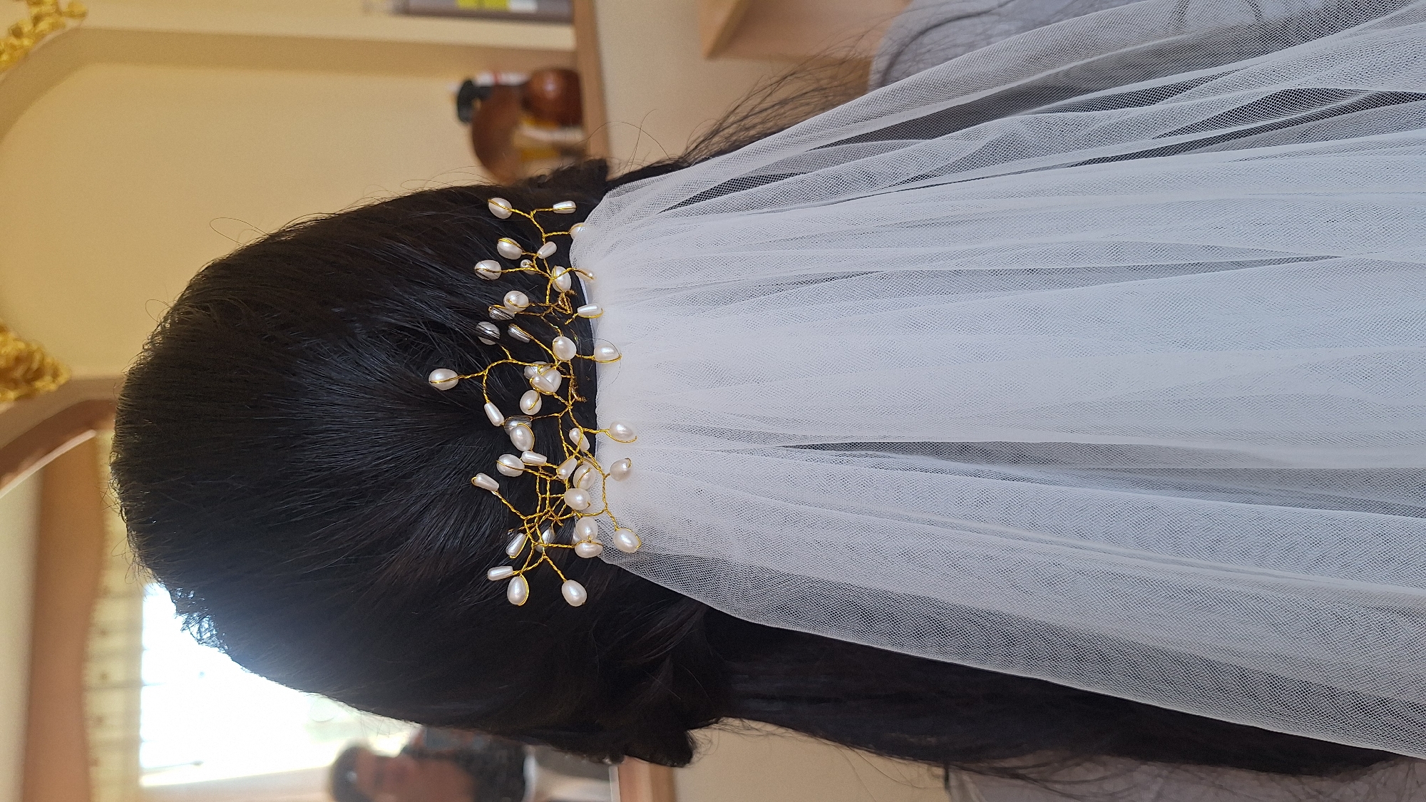 The image shows the back of a person's head, adorned with a wedding veil and a pearl hairpiece. The hair is dark and styled in braids. The veil is white and sheer, cascading down the back. The hairpiece is a delicate arrangement of gold wire intertwined with small, white pearls. The background is out of focus, but appears to be an indoor setting with a warm-toned wall and a window with sunlight streaming through. The overall impression is that of a bridal preparation. The location in Ogre, Latvia, is not discernible from the image.