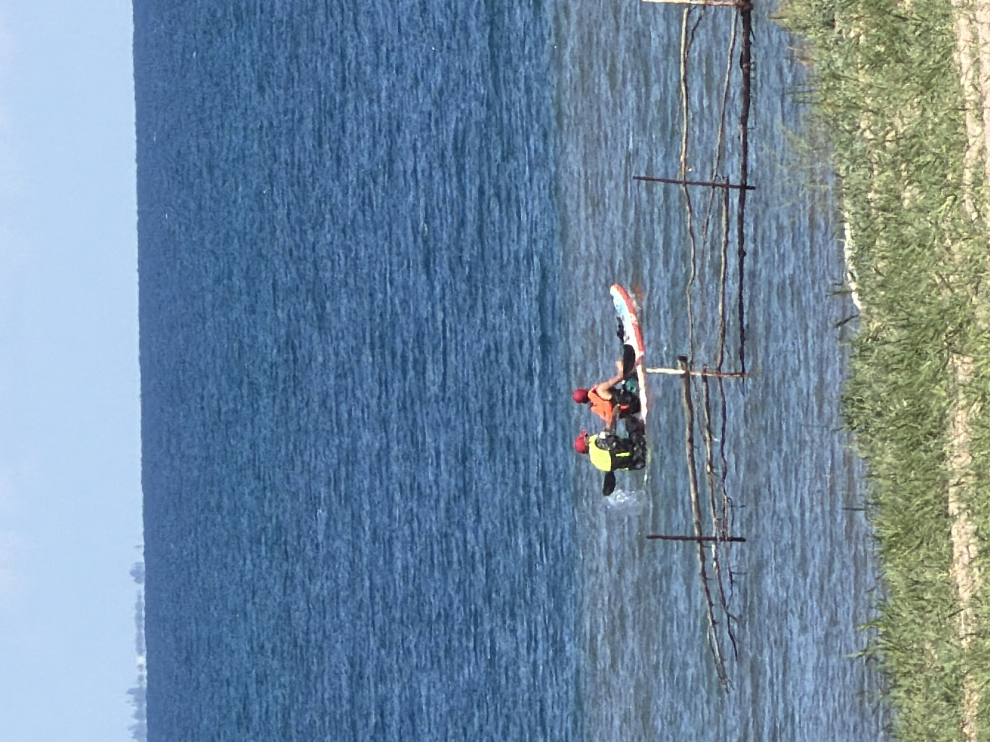 Two individuals, each wearing a red cap and a life vest, are in a kayak on the ocean. One person, on the left, is using a paddle. The kayak is white with red accents. The ocean is blue with moderate waves. A thin fence made of branches is visible in the foreground, partially submerged in the water. Beyond the water, a distant coastline with a few trees is visible on the horizon. The sky is clear and blue.