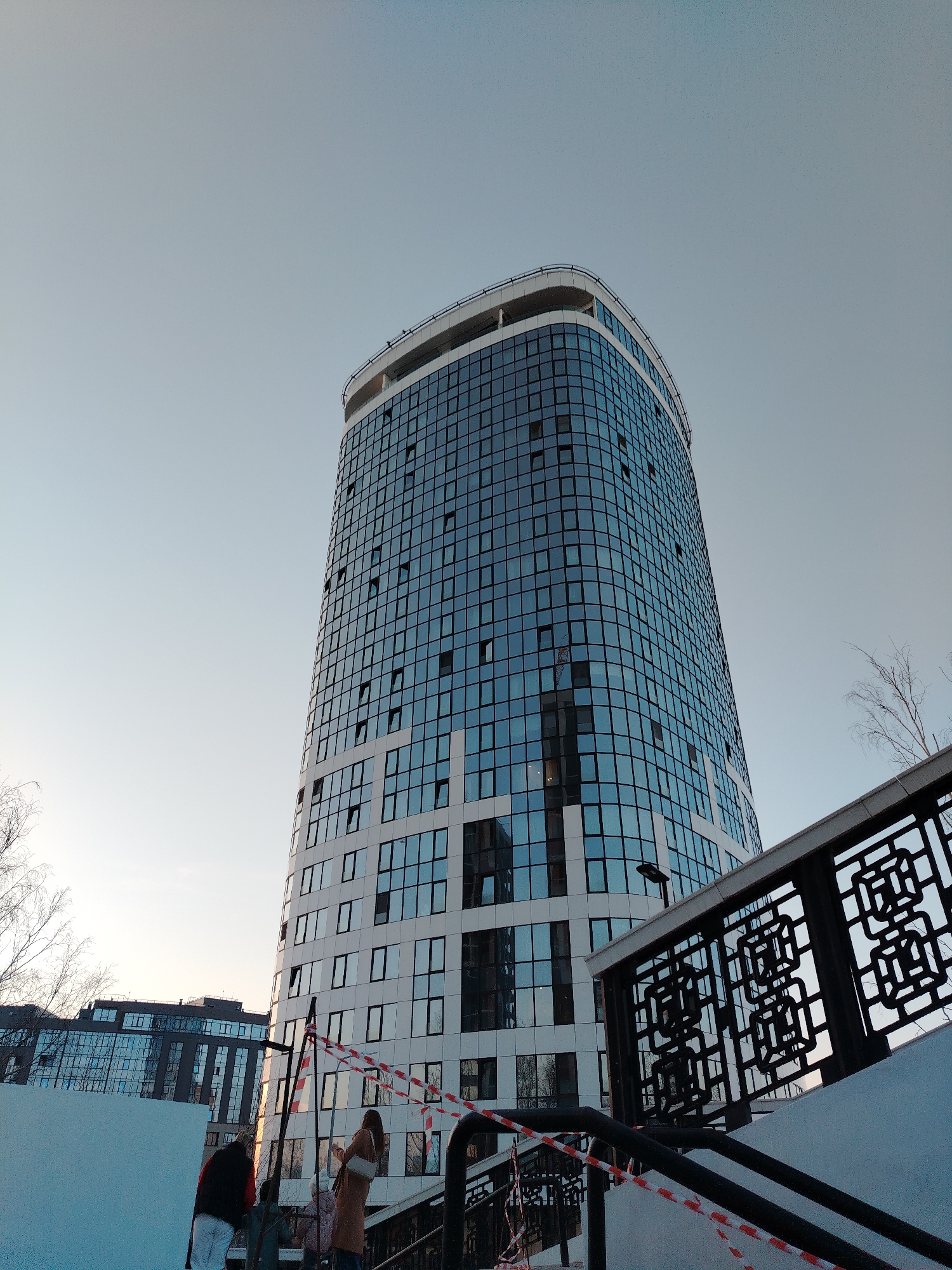 The image depicts the Izhevsk Plaza business center, also known as Respublika, located at 19 Pushkinskaya Street in Izhevsk, Udmurt Republic, Russia. The primary subject is a tall, modern, cylindrical high-rise building with a predominantly blue-tinted glass and white panel facade, curving gently upward to a rounded, white rooftop structure. Numerous windows are visible, some partially open.
In the foreground, a black metal railing with intricate, geometric cut-out patterns ascends from the lower right. Below this railing, a section of outdoor stairs is present, partially obscured. Red and white striped barrier tape, typical for hazard or construction zones, is draped across the lower part of the stairs and a black metal support structure. A plain white wall or surface is visible in the lower left quadrant.
Several individuals are present near the base of the stairs and the white wall. One person, seen from the back, wears a dark vest over a red garment. Another individual, also viewed from the back, wears a light brown coat and carries a white shoulder bag, appearing to look towards the main building. Other indistinct figures are partially visible nearby.
The sky is a clear, light blue, indicating daylight. Bare tree branches are visible on the left and right edges, suggesting a dormant season. In the background, to the left of the main high-rise, other contemporary buildings with glass facades are discernible. The perspective is low-angle, looking up at the central building.