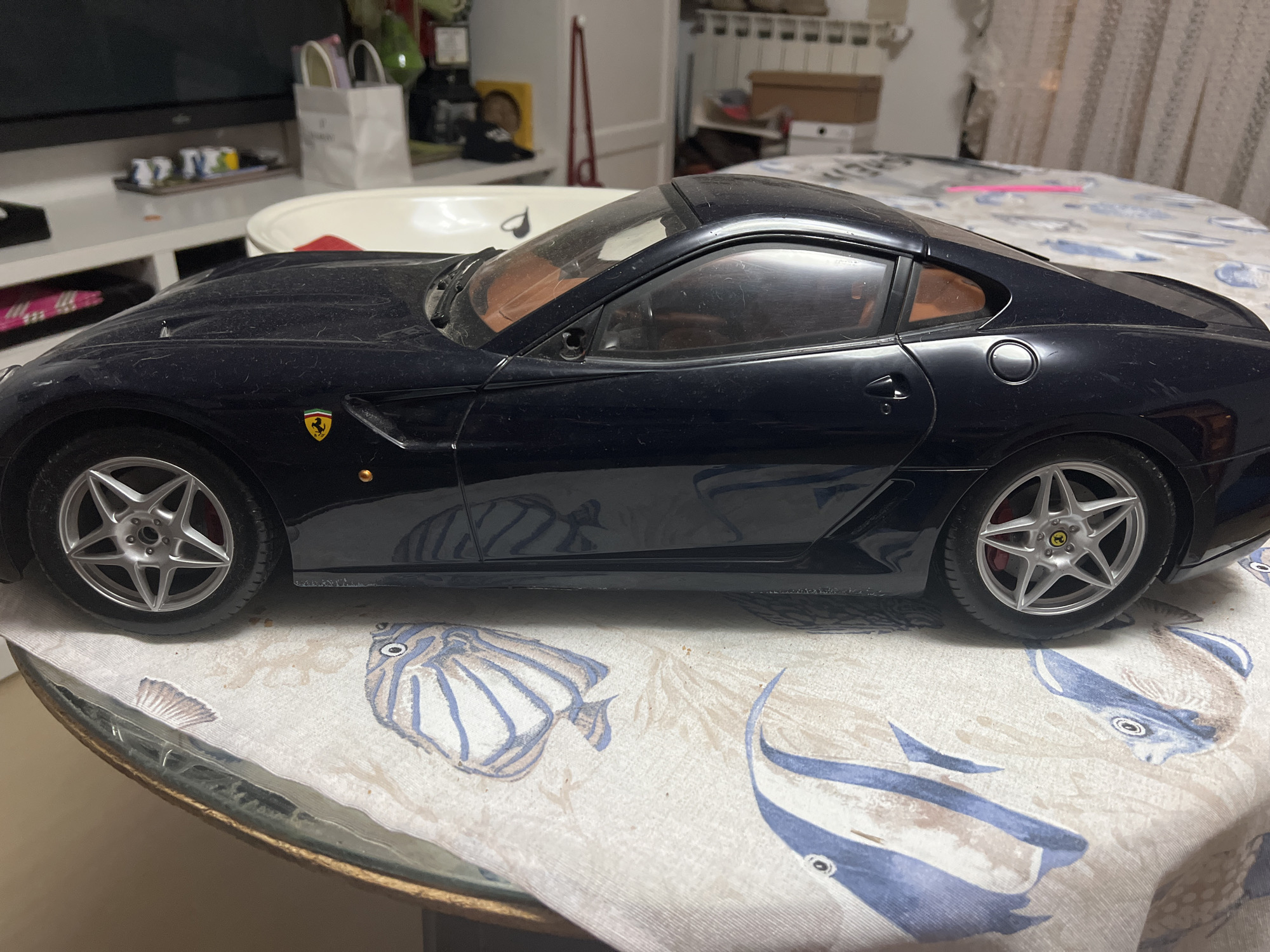 The image shows a close-up, side-profile view of a dark blue model Ferrari, possibly a 599 GTO, resting on a round table covered with a cloth patterned with illustrations of fish and seashells. The car is dusty. The interior appears to be tan leather. The car has silver five-spoke wheels with a yellow Ferrari emblem on the center caps, and red brake calipers are visible. In the background, shelves are stocked with items including boxes and bags, and a television screen is visible. The scene suggests an indoor setting, possibly a home. No people are visible in the image. The location is not explicitly stated to be Acquaviva, Italy, and no visual cues in the image suggest this specific location.