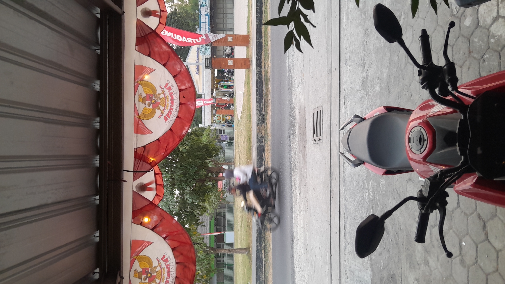 The image shows a street scene in Balun, Indonesia. In the foreground, the top of a red motorcycle is visible. Beyond the motorcycle, a street runs horizontally across the image. On the far side of the street, there are buildings and trees. Red and white banners with the Garuda Pancasila symbol are draped across the top of the scene. A motorcycle with two people on it is moving from left to right in the middle ground. The lighting suggests it is dusk or dawn.