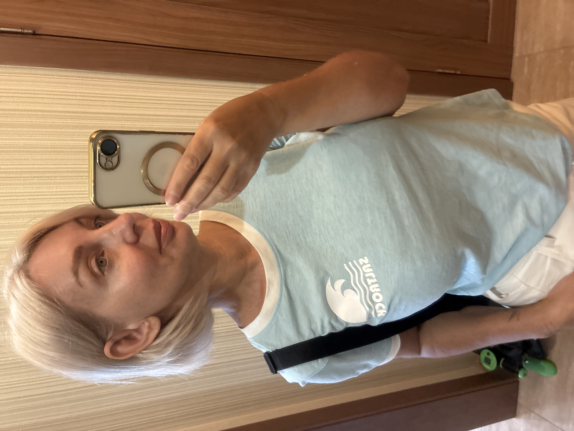 A woman with short blonde hair is taking a mirror selfie in a room with striped wallpaper and a wooden door. She is wearing a light blue t-shirt with a white ringer neckline and white shorts. A black strap from a bag is visible over her left shoulder. She is holding a smartphone with a gold-rimmed case and a circular attachment in her right hand. A small green toy is visible on the floor to the left. The image appears to have been taken indoors, likely in an apartment or hotel room. There is no specific indication of the location being in the city of Sochi, Russia.