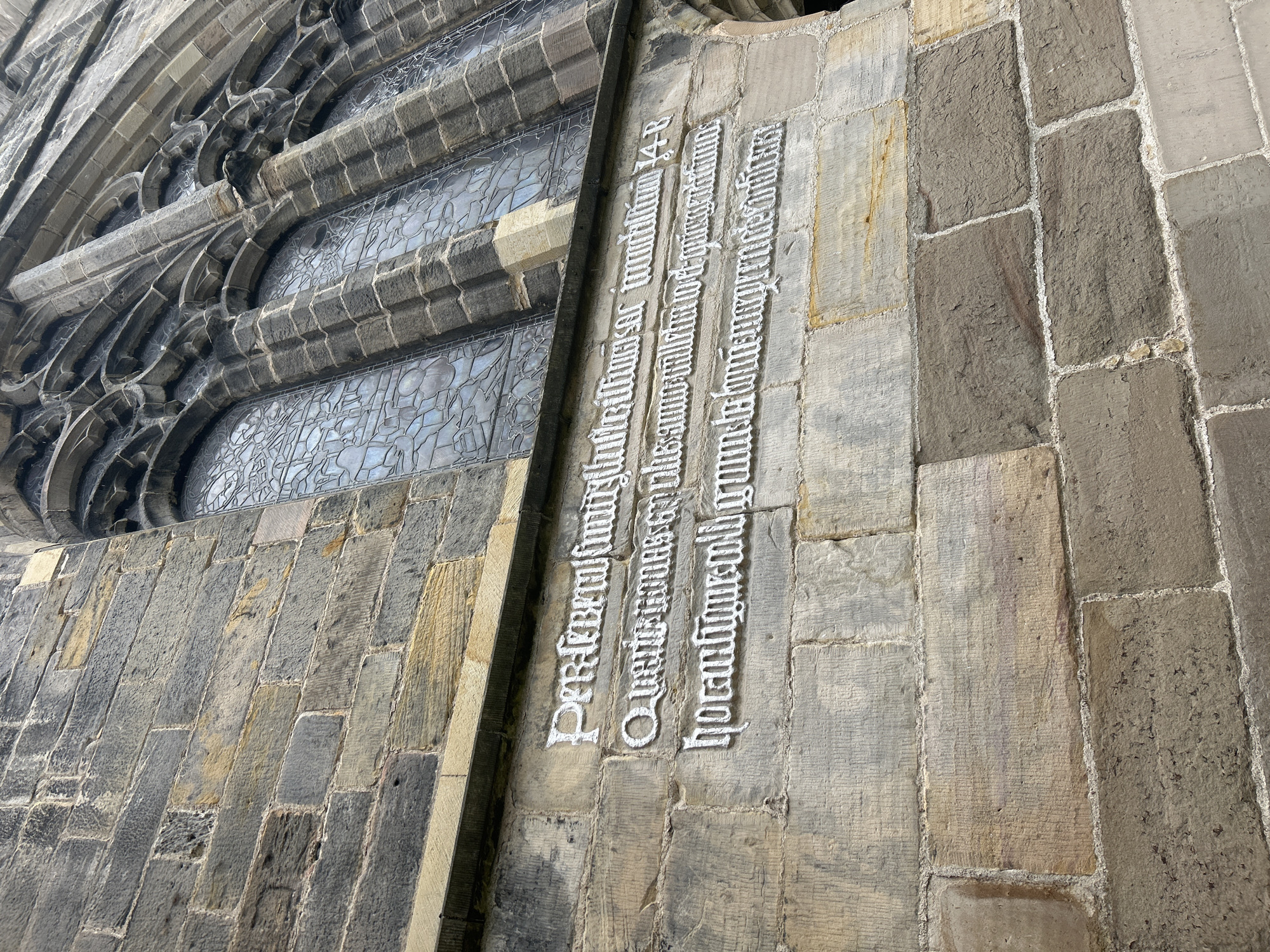 This image depicts a section of a building constructed from stone blocks, likely a church or similar historical structure, located in the city of Aberdeen City, United Kingdom. A window with intricate tracery is visible on the left side of the image. To the right, inscriptions are carved into the stone facade in a gothic style font. The first line appears to end with "multum 14-B", while the other lines are difficult to discern due to the font. The lighting suggests it is daytime, and the overall environment suggests an old and well-maintained building. There are no people visible in the image.