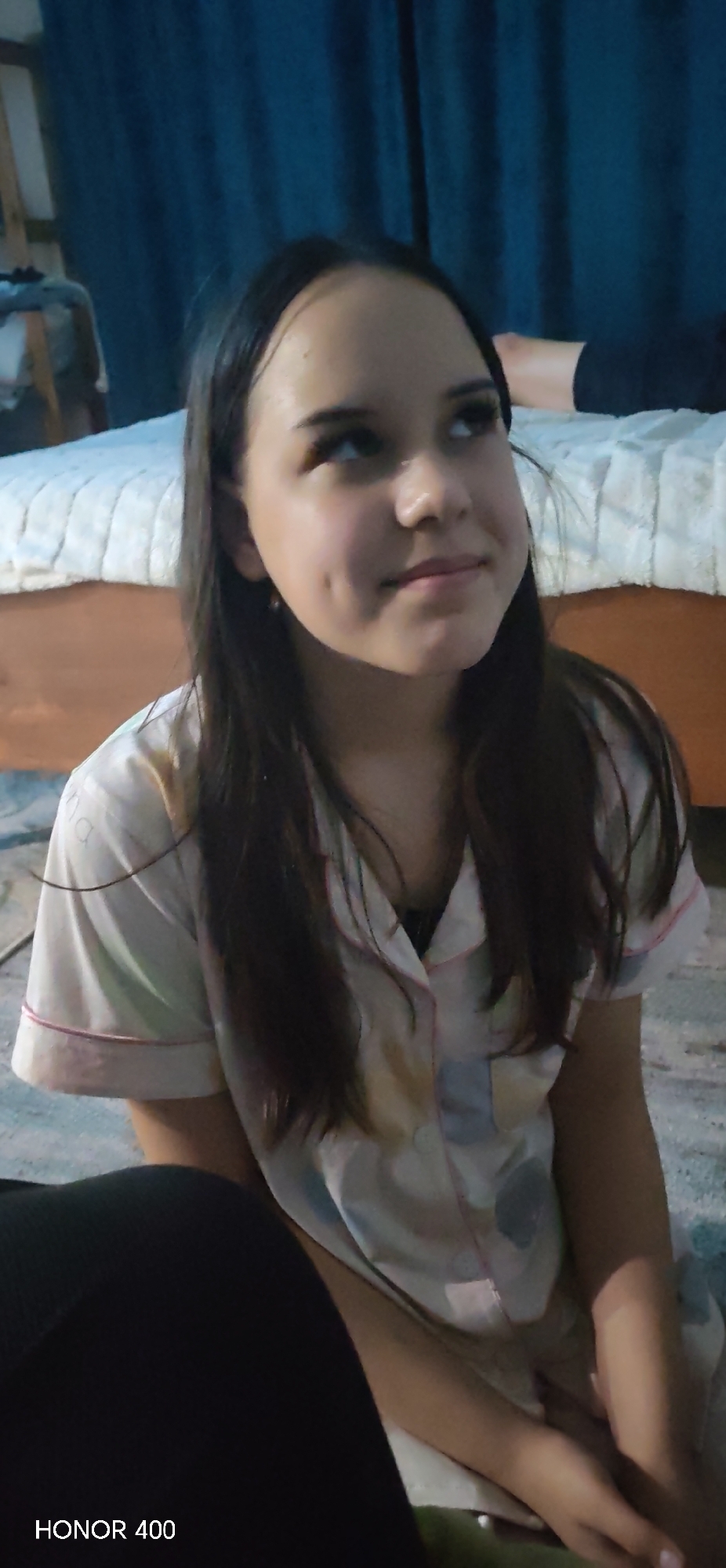 A young woman with long, dark hair and light skin is seated on the floor, looking upward and to the right. She is wearing what appears to be a light-colored patterned pajama top. Her face is visible and shows dimples. A bed with a white, textured bedspread and wooden frame is visible in the background, along with a dark blue curtain. Part of a person's leg, covered in black fabric, is visible on the bed. A black object is partially visible in the lower left foreground. The bottom left corner displays the text "HONOR 400." The location is most likely indoors, and there is no information in the image that identifies the city as Noviy, Russia.