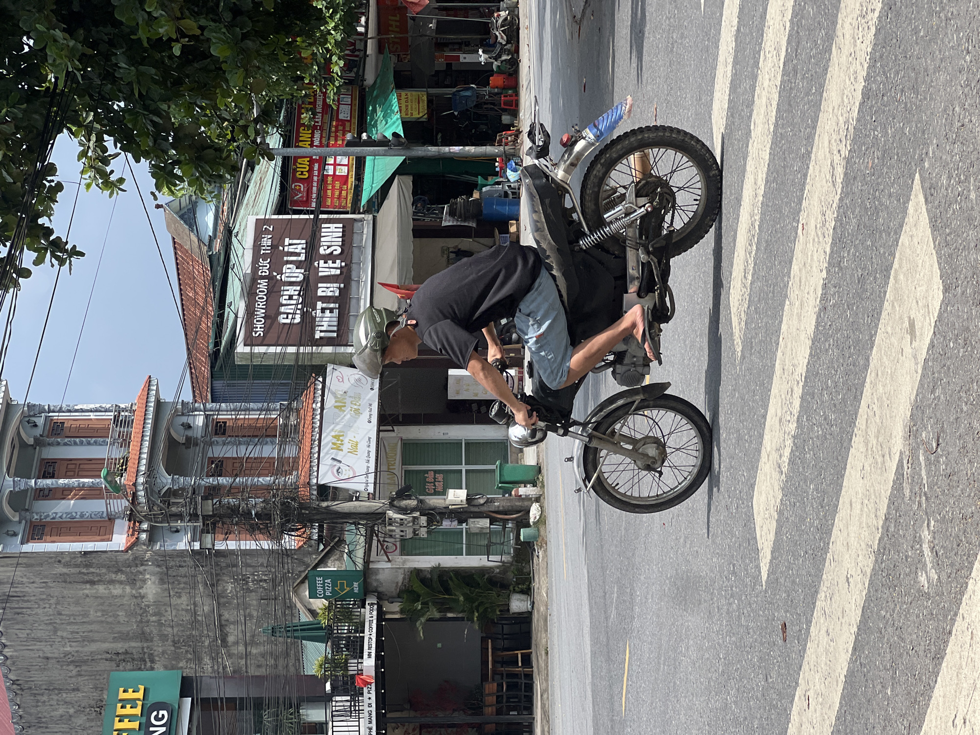 In Tân Quang Commune, Vietnam, a man wearing a light-colored helmet and a black t-shirt is riding a motorcycle with a gray seat and a black fender across a pedestrian crossing. He is wearing light blue shorts and flip-flops. The motorcycle has a silver exhaust pipe and a blue and white object attached to the rear fender. The background shows a street with buildings, trees, and utility poles. One building has a sign that says "COFFEE PIZZA" with an arrow pointing to the right. Another sign displays "SHOWROOM ĐỨC THỊN 2 GẠCH ỐP LÁT THIẾT BỊ VỆ SINH". The sky is overcast.