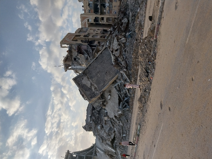 The image captures a scene of widespread destruction in what appears to be a residential or urban area, presented with a 90-degree counter-clockwise rotation. When viewed upright, the scene depicts a vast expanse of rubble and severely damaged buildings under a partly cloudy sky, with a road running alongside the devastation.
On the left side of the frame, a massive amount of debris dominates, consisting of broken concrete, twisted rebar, and pulverized building materials. Several multi-story buildings are either partially collapsed or reduced to skeletal frames, with floors and walls blown out, exposing interiors. A prominent, large concrete slab is tilted dramatically, resting precariously amidst the rubble. In the background, more damaged structures extend into the distance, hinting at the extensive scale of the destruction.
To the right, a paved road stretches across the lower portion of the image. It appears to be an asphalt surface, showing some signs of wear and dust. The road runs parallel to the area of destruction, separated by a rough embankment of dirt and smaller debris.
Several figures are visible near the edge of the rubble and the road. One individual, dressed in light-colored clothing (possibly pink or beige), stands in the mid-ground, close to the debris, appearing to observe the scene or simply walking past. Further in the distance, another person in darker attire can be seen near the rubble. A third figure is partially visible in the foreground, also positioned near the destruction.
The sky, visible in the upper-left section of the upright image, is partly cloudy. Patches of blue sky are interspersed with white, cumulus clouds, suggesting either an early morning or late afternoon light, as indicated by the soft illumination and lack of harsh shadows. The overall impression is one of profound devastation and loss. No discernible text is visible in the image.