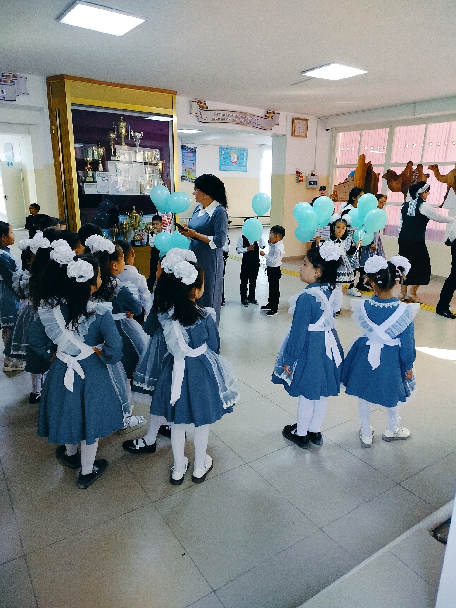 The image depicts a scene inside what appears to be a school hallway in the city of Kölkent, Kazakhstan. A group of young girls, dressed in blue school uniforms with white accents and large white bows in their hair, are gathered in the foreground. They are facing away from the camera, towards the center of the hallway. In the background, a woman in a blue uniform, likely a teacher, holds a mobile phone and stands near a cluster of light blue balloons. Several other children, boys in black suits and white shirts, are also visible in the mid-ground, some holding balloons. The hallway features a large display case filled with trophies and certificates. A clock is visible on the wall, along with decorative elements and windows with pink coverings. The floor is tiled. The overall impression is a school event, possibly a celebration or ceremony, with children dressed formally and participating in activities.