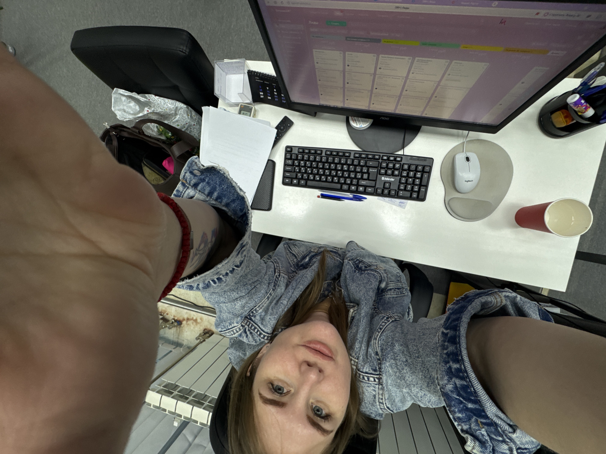 The photograph, taken from a high angle, shows a woman in a denim jacket at a desk with a computer. The desktop displays a Kanban-style project management tool. A keyboard, mouse, pen, paper, calculator, calendar, and cup are on the desk. A pen holder sits on the right edge of the desk. A purse rests on a chair behind the woman. A radiator is visible in the background. This scene is possibly in Perm, Russia, but there is no indication of a specific location to verify that statement.