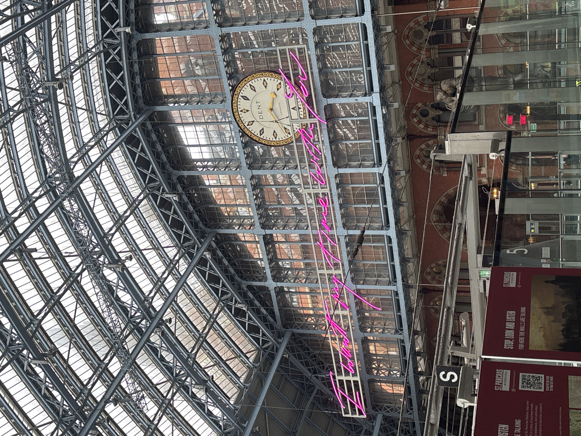 This interior shot shows the ornate, iron-and-glass ceiling structure of St. Pancras International railway station in London, United Kingdom. A prominent clock with "DENT LONDON" inscribed on its face hangs suspended from the ceiling. A neon art installation spelling out a phrase extends diagonally below the clock. The station's upper walls are visible behind the glass. Signage including the text "STOP, LOOK AND LISTEN FOR HERE THE WALLS ARE TALKING" and the station name "ST. PANCRAS INTERNATIONAL" is visible. A platform number "5" is visible in the upper right.