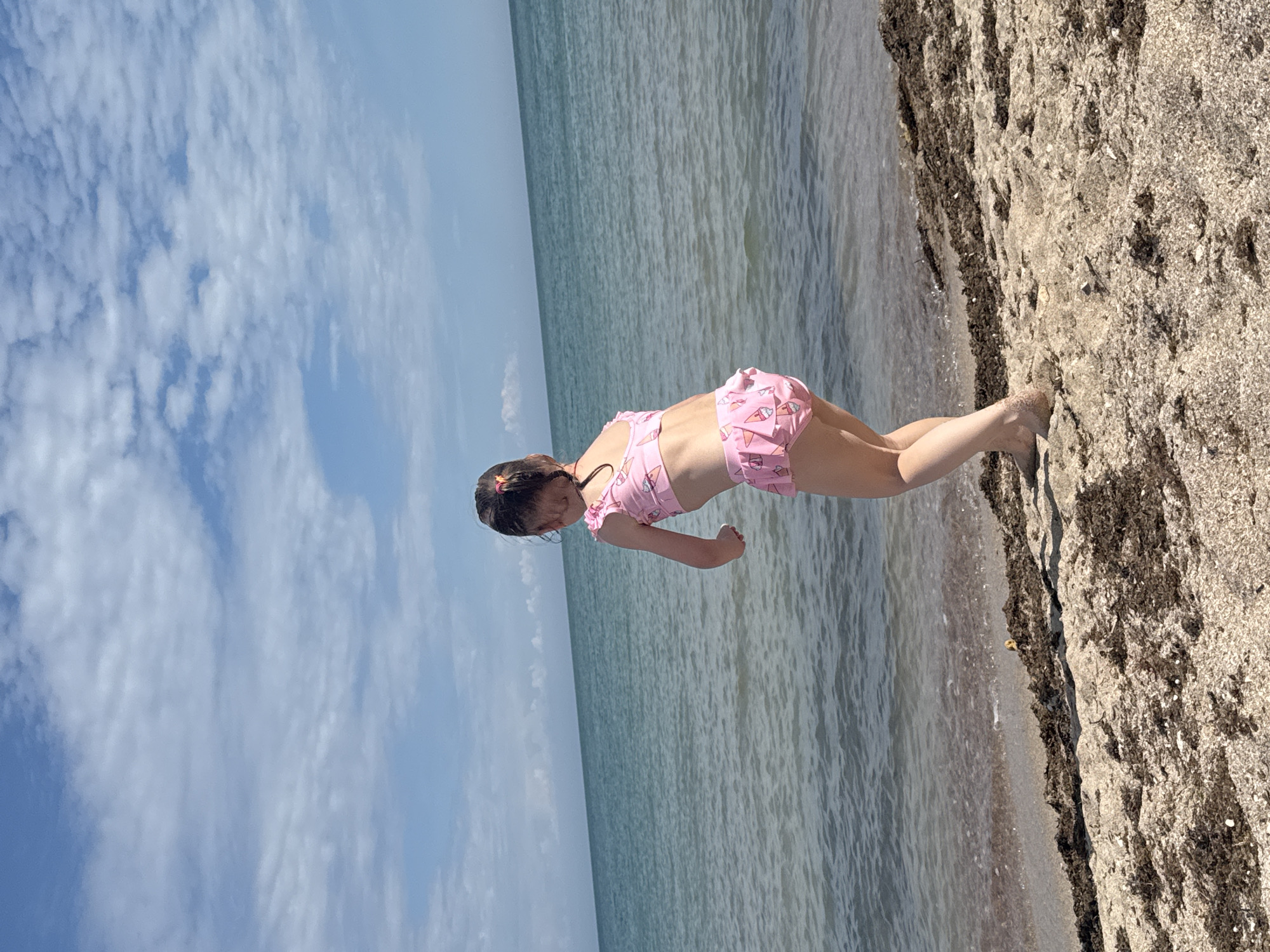 A young girl in a pink swimsuit with an ice cream pattern is standing on a sandy beach next to the sea. The sky is blue with scattered white clouds. The location is likely in or near Yevpatoriya, Ukraine, given the prompt's request.