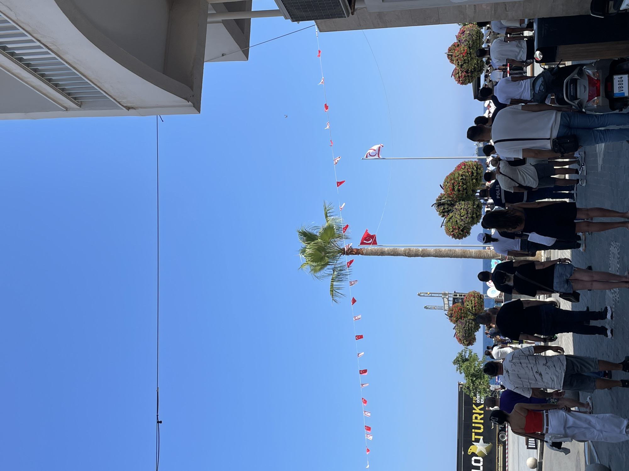 The image depicts a bustling street scene in Kyrenia, Northern Cyprus, on a clear, sunny day. A group of people are walking away from the camera, some of whom are wearing casual summer clothing. In the background, a palm tree stands tall, adorned with a string of small Turkish and Cypriot flags. A larger Turkish flag is also visible flying on a flagpole. To the right, a modern building with an arched entrance and balcony overlooks the street. To the left, a large truck with "YOLO TURK" printed on its side is parked. The street is paved, and there are decorative bushes with red flowers along the pathway. A motorcycle is parked on the right side of the frame. The overall impression is one of a lively public space, possibly a promenade or a town square.