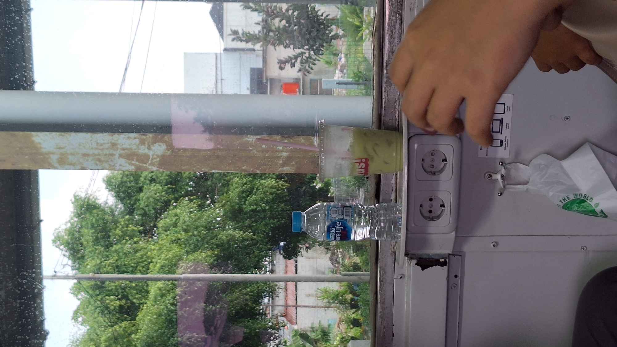 A person's hands are in the foreground, reaching towards electrical outlets. The background shows a window with trees and buildings visible outside, suggesting an urban or semi-urban environment. A bottle of water and a cup of green drink are placed on a shelf in front of the window. The outlets are mounted on a white panel. The overall scene appears to be inside a vehicle or a building with a window view. The mention of Karawang, Indonesia, is not directly supported by visual evidence in the image itself, but could be inferred from external context or metadata.
