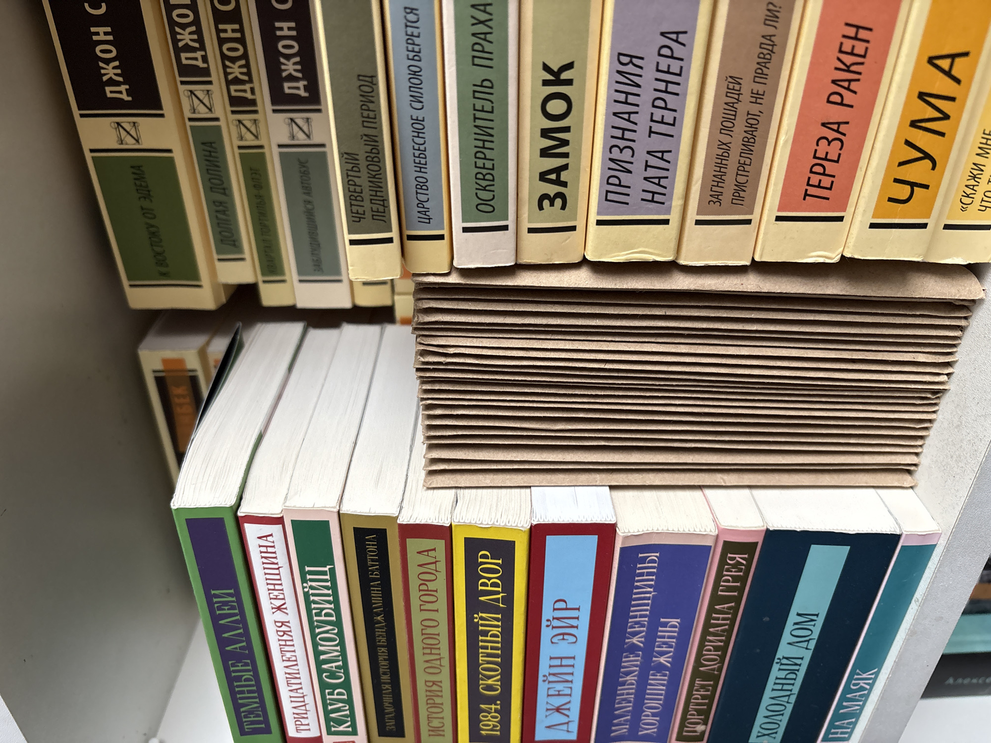 The image shows a shelf of books, likely in a bookstore or library. Most of the books appear to be written in Russian, with titles such as "To the East of Eden" and "Dark Alleys" visible on the spines. The shelf is densely packed, with some books stacked horizontally on top of others. The environment seems enclosed and focused on literature.