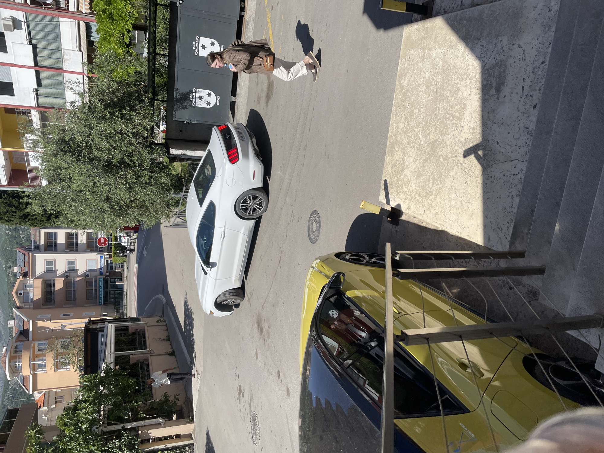 The image captures a street scene in Budva, Montenegro, showing a white and a yellow car parked alongside a road in front of a building. A woman is walking down the road near the white car. The building features a sign written in the local language, alongside trees and urban surroundings. In the background, a glimpse of Budva's architecture is visible, with residential and commercial buildings lining the street.
