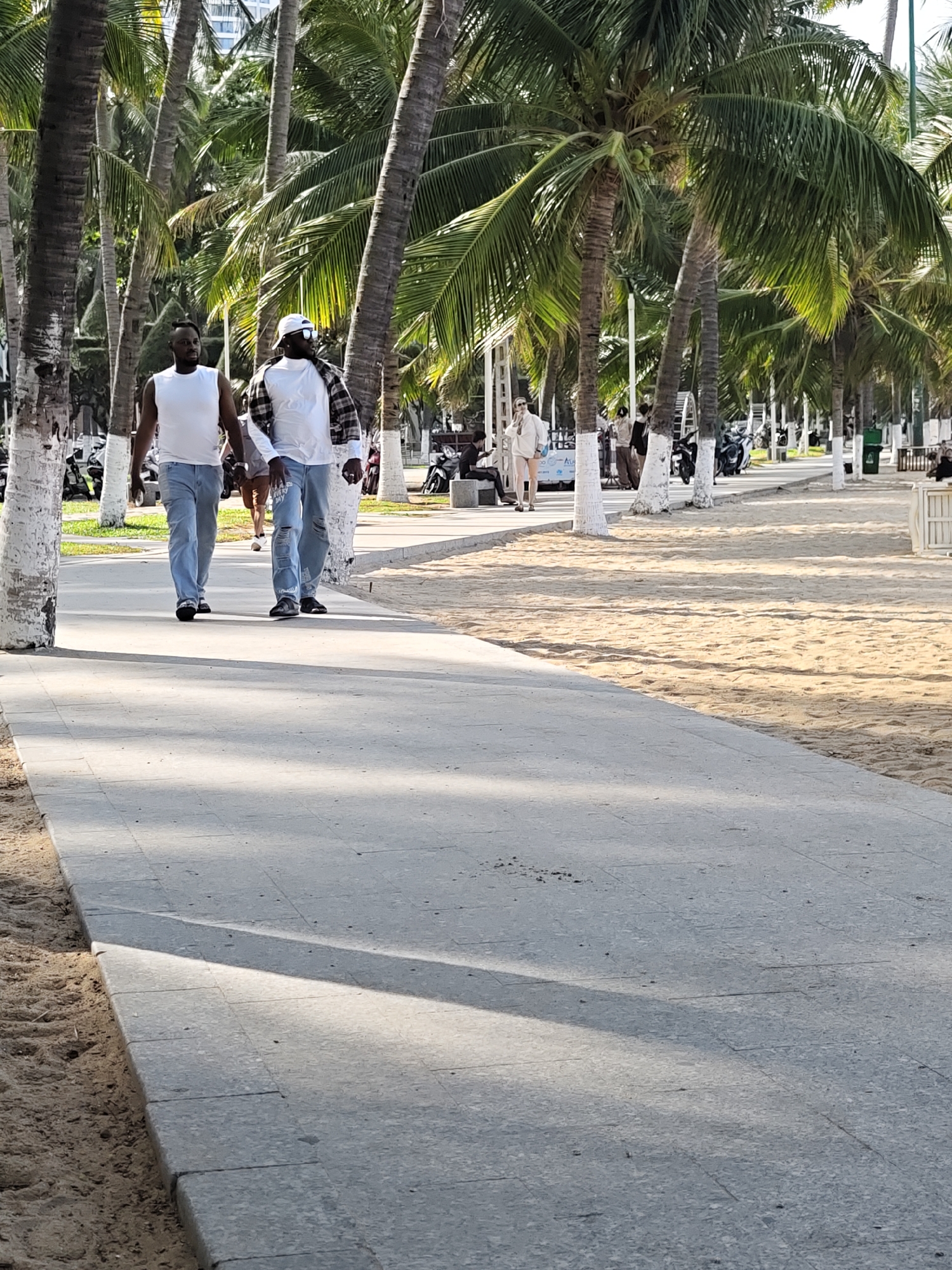 Two men are walking on a paved pathway next to a sandy beach lined with palm trees. The man on the left wears a white sleeveless shirt and blue jeans. The man on the right wears a white t-shirt, blue jeans, and a plaid shirt tied around his waist, and a white cap. In the background, other people, motorcycles, and white-painted trees are visible. The scene is likely located in Nha Trang Ward, Vietnam, suggested by the presence of palm trees and a beach setting common to coastal tourist areas in Vietnam.