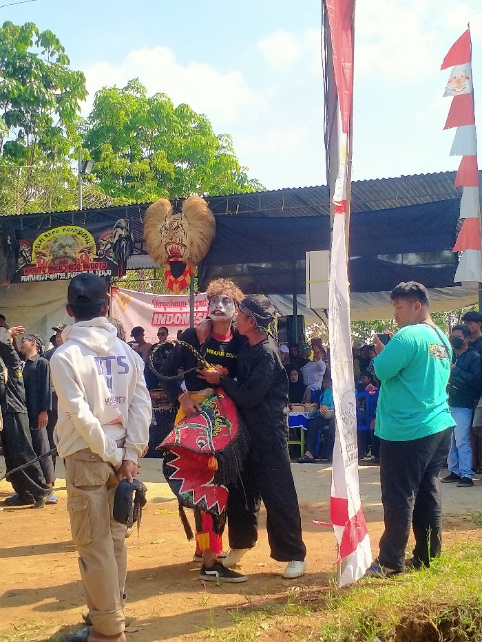 An outdoor cultural performance is captured in Plosorejo, Indonesia, specifically in Wates, Kebumen, as indicated on a background banner. The scene is set on a dirt ground under bright daylight, with a crowd of spectators observing. In the midground, a central performer with white and black theatrical face paint, curly hair, a black t-shirt (partially reading "IMAN EDUK"), and red/yellow patterned trousers is being supported by an individual dressed in black with a dark head covering. The supporting individual has their arm around the performer's shoulder and holds a red, black, and white decorative prop, possibly a hobby horse associated with Jathilan. The performer holds a black braided whip or rope. To the right, another individual in a teal t-shirt records the event with a horizontal smartphone. In the background, a temporary stage structure with a corrugated metal roof is draped with black fabric. Mounted centrally on this structure is a large, ornate tiger-like mask with prominent fur/feathers. Banners include "SENI REOG TRADISIONAL SINGO KEDU BUDOYO" and "DIRGAHAYU REPUBLIK INDONESIA." Lush green trees are visible against a clear blue sky above the stage. In the foreground, a vertical pole draped with red and white fabric stands on the right, and a person in a white hoodie, black cap, and khaki pants stands with their back to the viewer on the left, holding a dark object behind them.