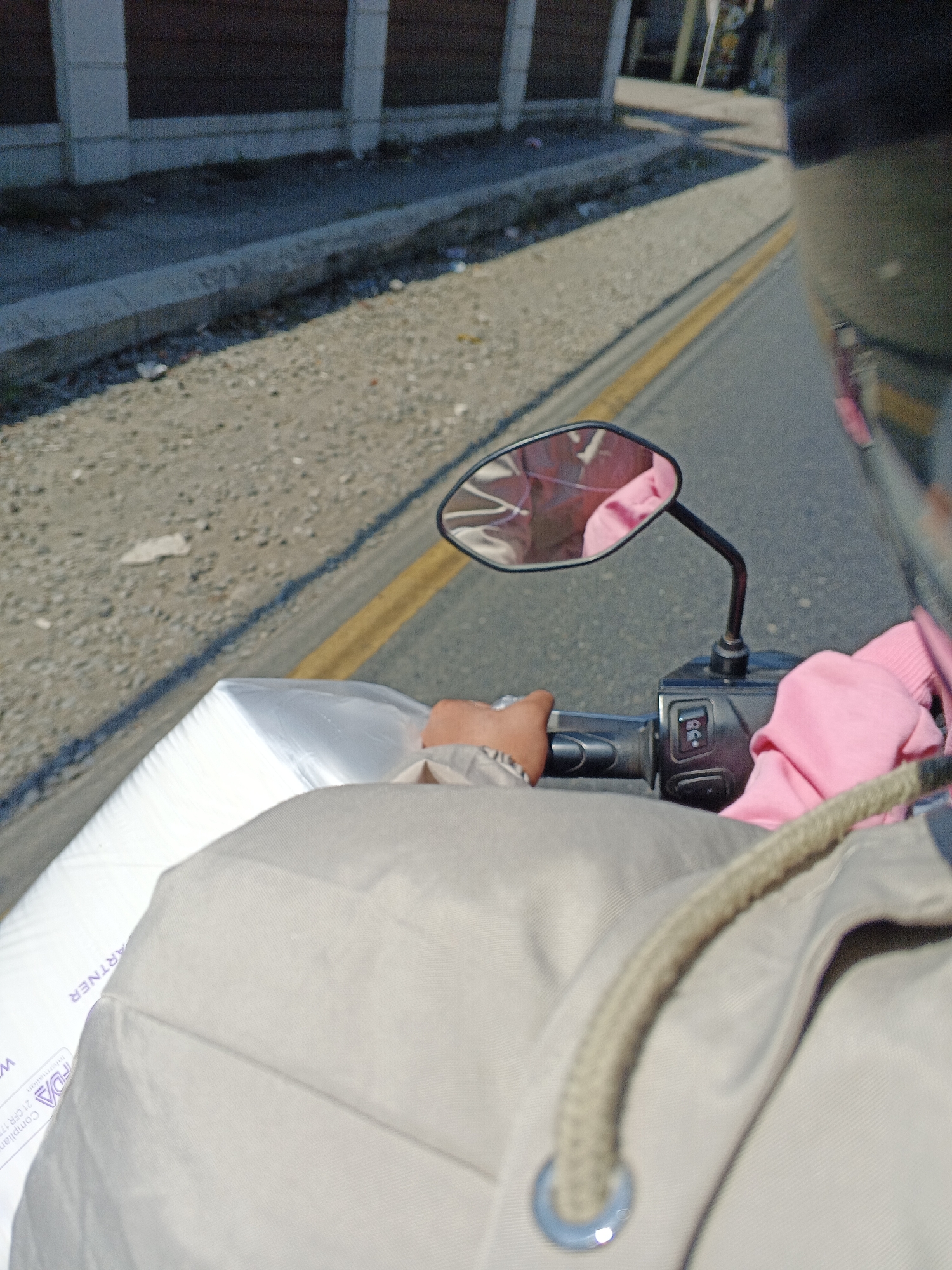 The image depicts a point-of-view shot from a person riding a scooter or motorcycle, likely on a street in Sukabumi, Indonesia. The rider's hands, wearing a light-colored jacket and holding the handlebars, are visible. A large white package, possibly wrapped in plastic, is secured in front of the rider. Pink fabric, likely clothing, is draped over the handlebars and on the rider's arm. The scooter's side mirror reflects a pink garment. The environment shows a paved road with a yellow line marking the lane, bordered by a gravel shoulder and a concrete curb. In the background, a dark fence and buildings are partially visible. The scene appears to be captured during daylight.