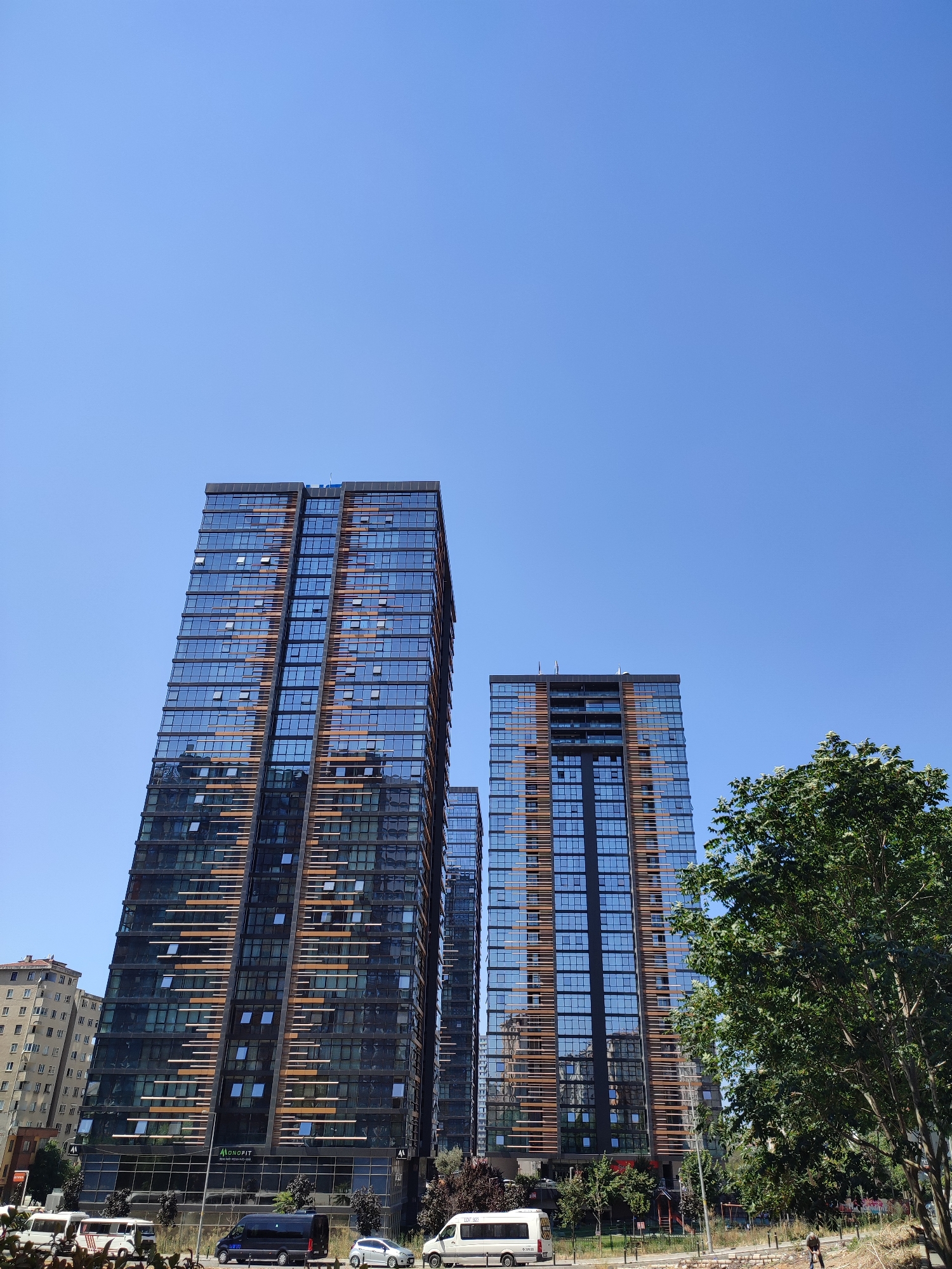 The image shows a street scene in Kadıköy, Turkey, on a clear, sunny day. Two modern, tall residential buildings with dark glass facades and vertical wooden accents dominate the center of the frame. The building on the left is taller and appears to be under construction or recently completed, with some windows open. The building on the right is slightly shorter and also features a similar glass and wood design.
In the foreground, a tree with green foliage is visible on the right side of the image. To the left of the modern buildings, an older, multi-story building with a lighter facade is partially visible. The street level is populated with several vehicles, including a white minivan, a dark minivan, a silver car, and a white van with text on its side. A few people are present on the sidewalk, with one individual walking away from the camera near the right edge of the frame. The sky is a bright, cloudless blue.
