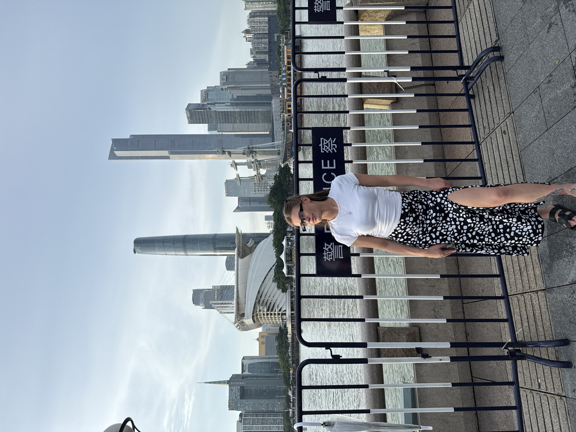 A young Caucasian woman with blonde hair pulled back in a ponytail, wearing glasses, a white t-shirt, and a black and white polka dot skirt with a high slit, stands facing right, looking to the left. She is positioned in front of a black and white striped barrier that separates her from a body of water, likely the Pearl River. Behind the water, a cityscape is visible with numerous modern skyscrapers, including the distinctive IFC tower and the Guangzhou Opera House. The sky is a pale blue with some clouds. The scene appears to be a waterfront promenade in Guangzhou, China, specifically in the Haizhu District, known for its modern architecture and riverfront views. The barrier suggests a restricted area, possibly for crowd control or safety along the waterfront. The woman holds her phone in her right hand.