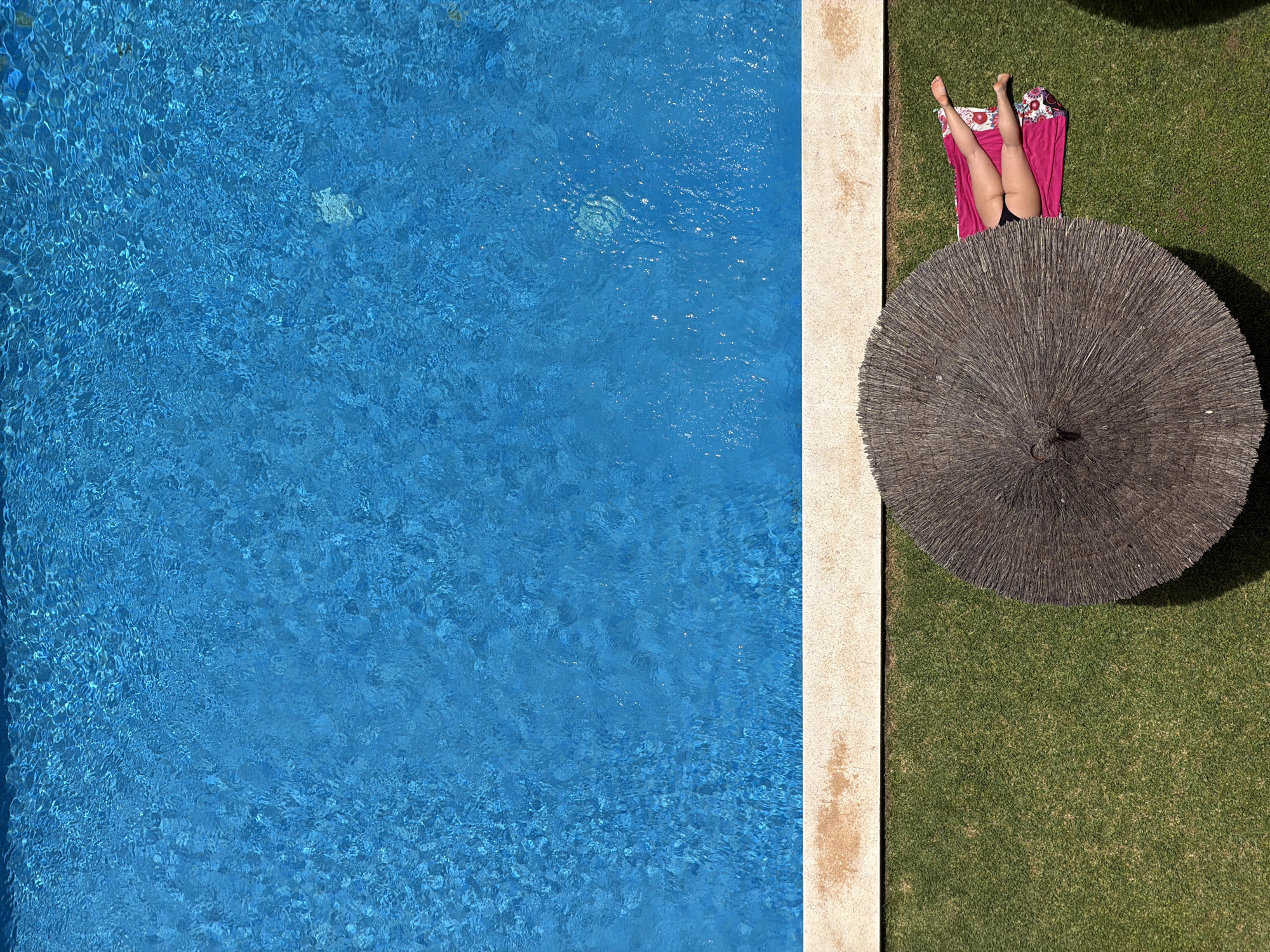 An aerial, high-angle full shot shows a person lying on a pink towel under a thatched umbrella next to a swimming pool. The person's legs, clad in black bikini bottoms, extend from under the umbrella towards the pool. The pool is filled with clear blue water and the person is on a patch of green grass. The edge of the pool is a light-colored concrete border.