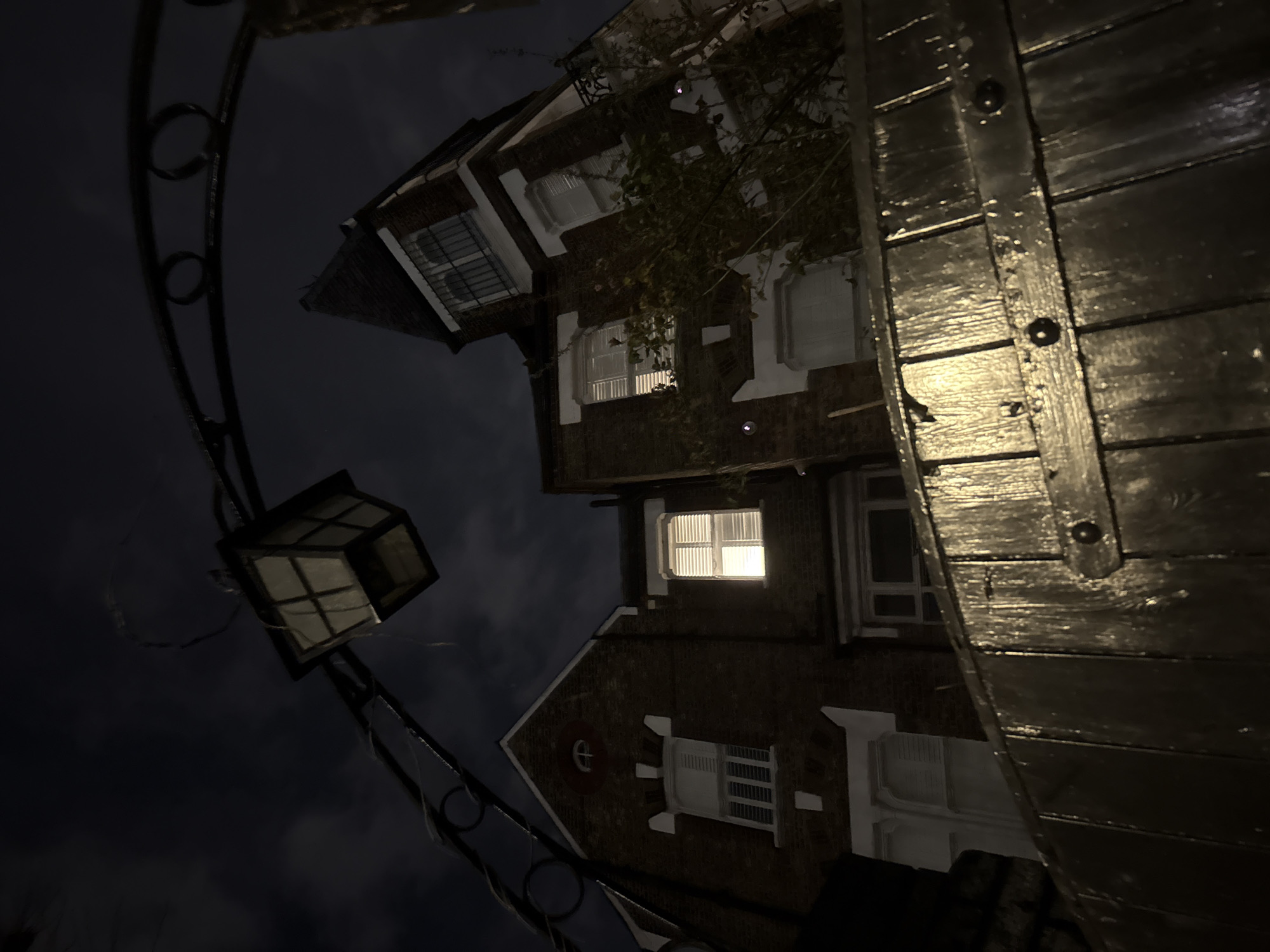 A low-angle, nighttime photograph depicts a multi-story residential building, likely in the City of London, United Kingdom, viewed from behind a dark wooden gate. In the immediate foreground, a dark, glossy wooden gate is present, composed of vertical planks secured by horizontal metal straps with visible rivets. Above the gate, a dark, ornate metal archway featuring circular and scrolled decorative elements extends across the upper frame. A traditional-style, dark lantern with visible panes hangs from this archway on the left side.
The main subject is a red brick building with white architectural trim around its windows and eaves, consistent with Victorian or Edwardian design. On the building's facade, a mid-level window in the left section is brightly illuminated from within, revealing closed white louvered shutters. Other windows on the building also feature closed white shutters. The leftmost section includes a gable with a small, oval-shaped window. The building's right side incorporates a turret-like structure topped with a dark roof and windows protected by dark metal grilles. Sparse, dark vegetation is visible clinging to parts of the building. The background consists of a dark, cloudy night sky. No individuals are present in the scene.