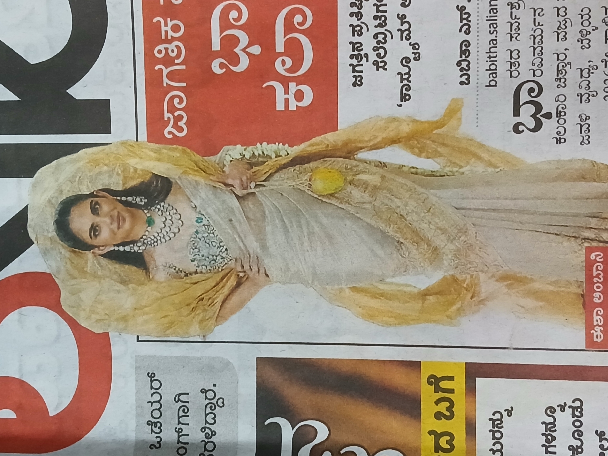 The image is a newspaper clipping featuring a woman in an elegant, light-colored sari, adorned with a necklace and draped with a golden shawl. She is the focus of an article titled “Jagatika V”, suggesting a global theme. The clipping includes text in Kannada script, mentioning figures like "Eesha Ambani" and “Babitha S. Salian,” possibly related to art, fashion, and celebrity culture. Text mentions “Kala Jagattina Pratishthi" which translates to world of art. The clipping also refers to “Ratha Ravivarma” and “Kalamkari Chittara,” indicating a connection to Indian art forms.