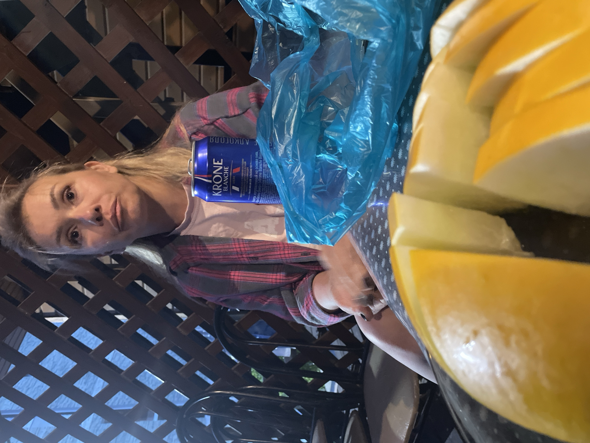 A Caucasian woman with blonde hair is sitting on a chair, facing forward. She is wearing a pink and grey plaid shirt over a pink shirt. A blue can of "Krone Blanche Biere" is on the table in front of her, next to a bright blue plastic bag. In the foreground, slices of melon are visible on a black surface. The background features a wooden lattice fence and dark chairs. The overall scene appears to be outdoors, possibly on a patio or deck.