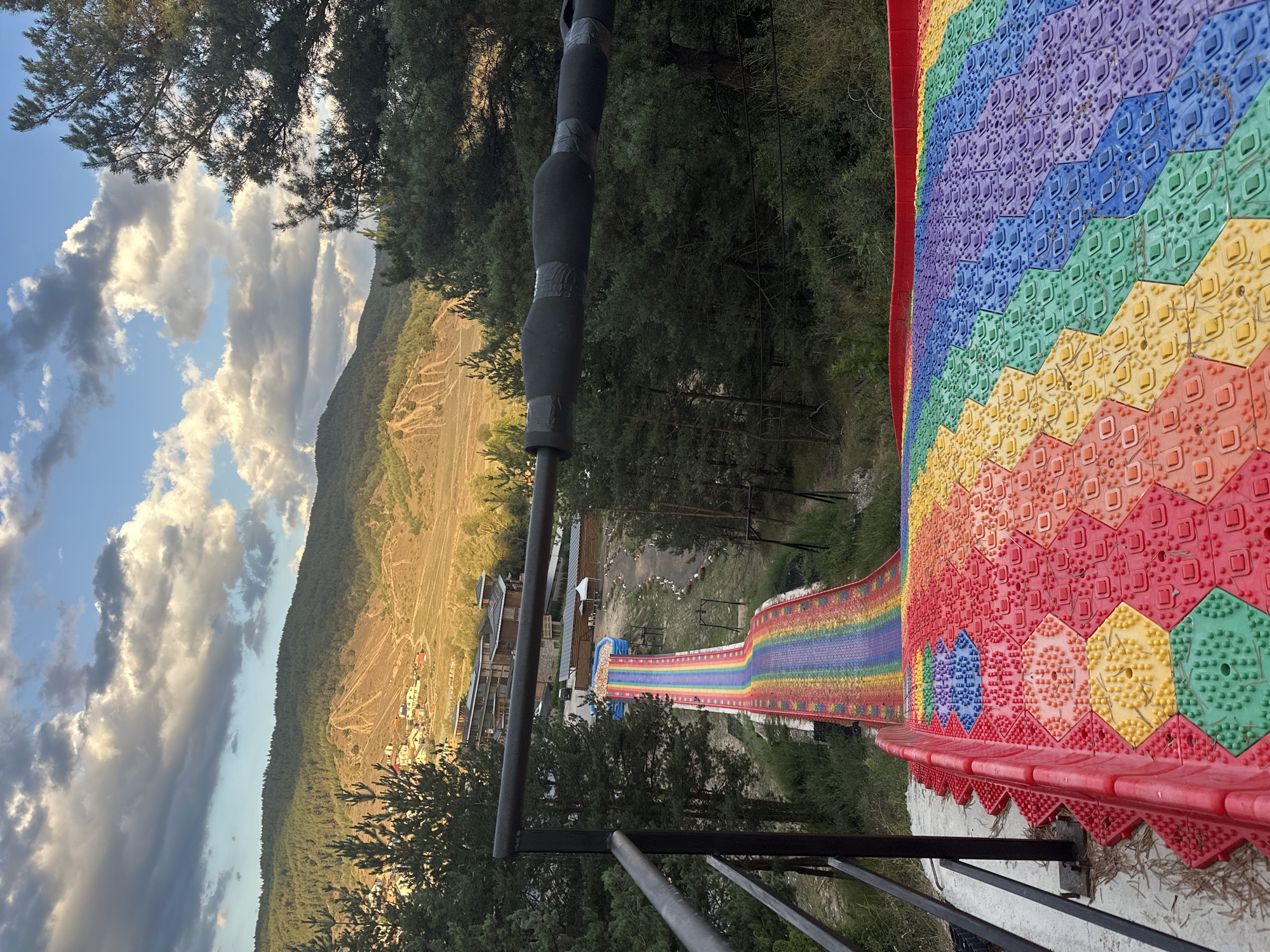 The image captures a vibrant, multi-colored slide resembling a rainbow, set against a backdrop of a lush green forest and rolling hills. The slide appears to be made of interlocking hexagonal tiles, each embossed with a pattern of small bumps, contributing to its unique texture. The slide starts with red and orange tiles at the forefront and transitions through yellow, green, blue, and purple as it extends down a slope. The surrounding area is covered in green grass and scattered pine trees. In the distance, the landscape features more trees and some buildings, suggesting a settlement or resort area. The sky above is partly cloudy, with dramatic formations of white and grey clouds illuminated by sunlight, hinting at either sunrise or sunset. A metal railing is visible on the left side of the image, indicating a viewing platform or the start of the slide. The scene is one of an outdoor recreational facility, likely designed for amusement. The image does not provide explicit details to confirm its location in Bania, Bulgaria, but it depicts a scenic and colorful attraction in a natural environment.
