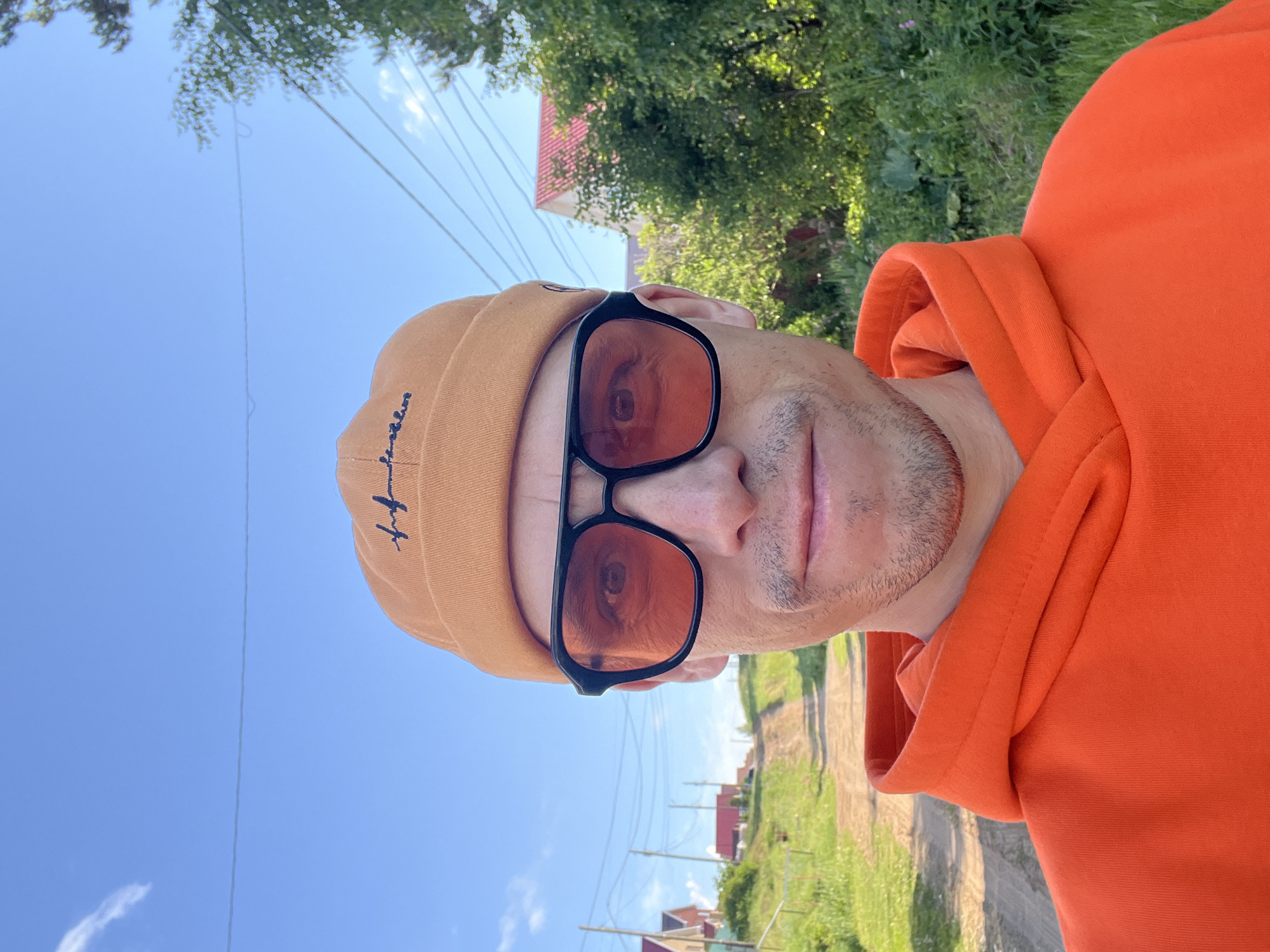 A person is captured in a vertically oriented close-up selfie, outdoors on a sunny day in what appears to be a rural or semi-rural setting in Chaikovsky, Russia. The individual, likely male, is wearing a light brown or tan beanie with dark, stylized text embroidered on the side, possibly a brand logo. He is also wearing black-framed sunglasses with reddish-brown tinted lenses, through which one of his eyes is partially visible, showing a reflection. Stubble is visible on his chin and upper lip. He is dressed in a bright orange hoodie.
The background features a clear blue sky with a few faint white clouds, indicating bright daylight. Lush green trees and foliage are visible, particularly in the upper left and right portions of the background. Distant houses with red roofs can be seen beneath the horizon, suggesting a settlement. Several thin wires, likely power lines or communication lines, stretch across the sky, with poles partially visible in the distance. A dirt road recedes into the background, flanked by grassy areas. The overall scene suggests a warm, pleasant day in a quiet, open environment.