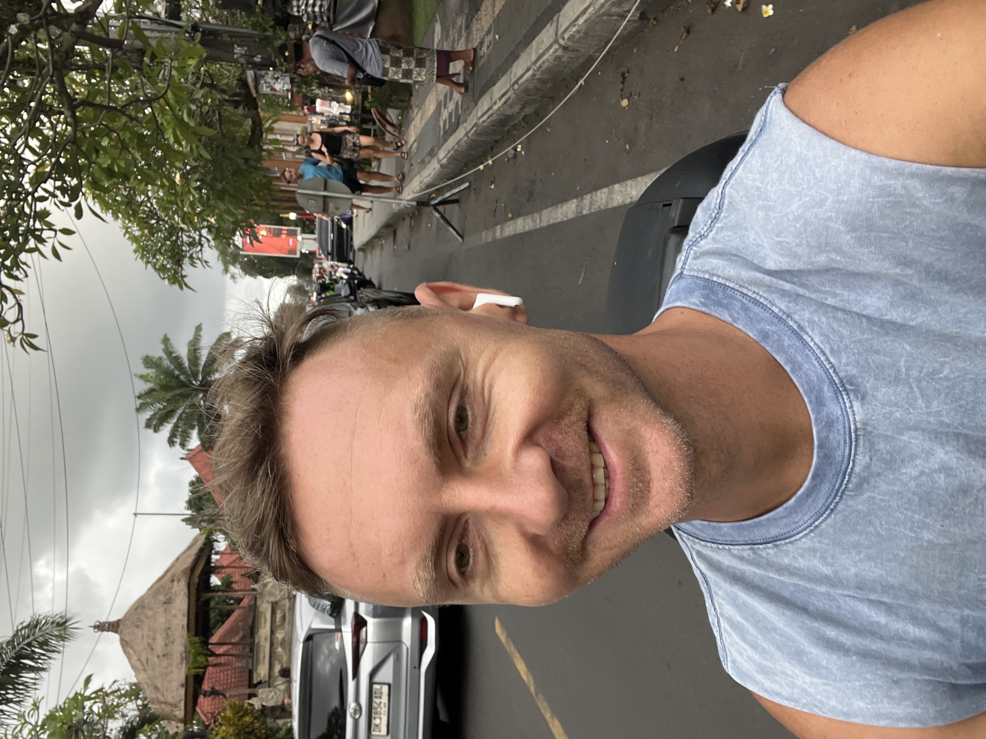 A close-up selfie shows a smiling man with light hair and a blue t-shirt, wearing a white wireless earbud. He is on a street in what appears to be Ubud District, Indonesia. In the background, there are parked cars, a traditional Balinese building with a thatched roof, palm trees, other people walking along the sidewalk, and various shops and establishments. The sky is overcast.