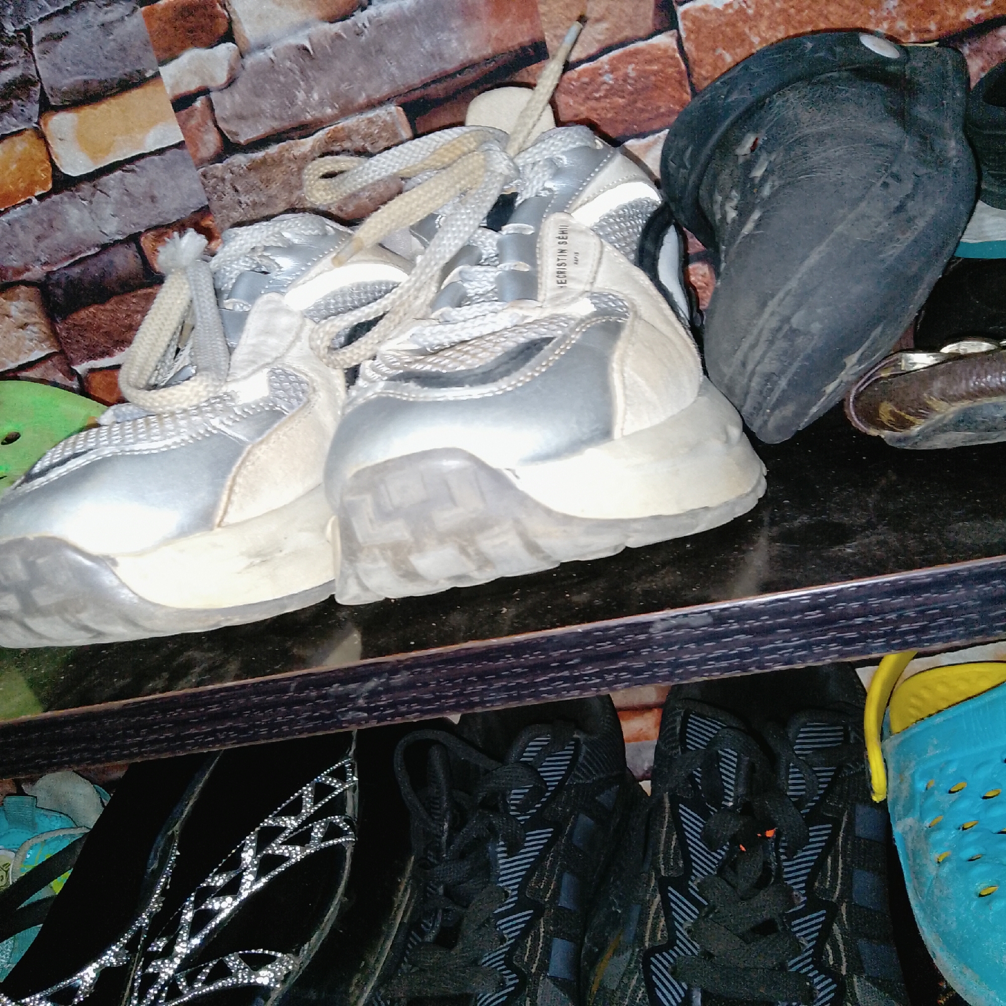 The image shows a shelf with several pairs of shoes. In the foreground, there are two pairs of athletic shoes, one white and silver, and another black with a striped pattern. To the right of the athletic shoes, a blue plastic clog with yellow trim is visible. Behind the athletic shoes, a pair of black boots is partially visible. The background consists of a brick-patterned wall. The lighting is dim, suggesting an indoor setting. The exact location in the city of Unknown, Unknown cannot be determined from the image.