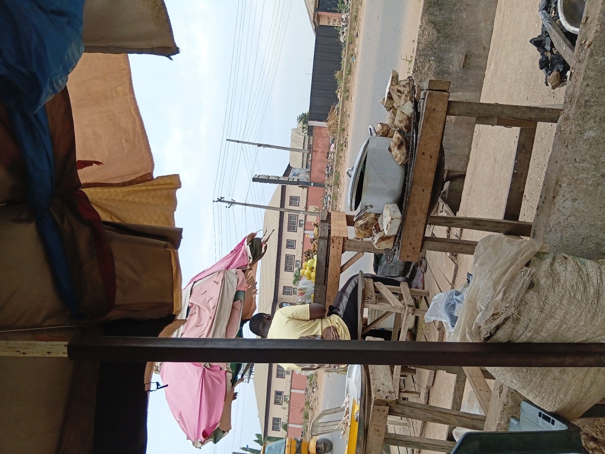 The image shows an outdoor market scene in Akute, Nigeria. A man in a yellow shirt sits at a wooden stall. The stall displays food items, including what appears to be boiled yam in a large pot. Bags and other objects are arranged around the stall. The stall is covered by a makeshift canopy of tarpaulins. A road and buildings are visible in the background. Overhead power lines are present.