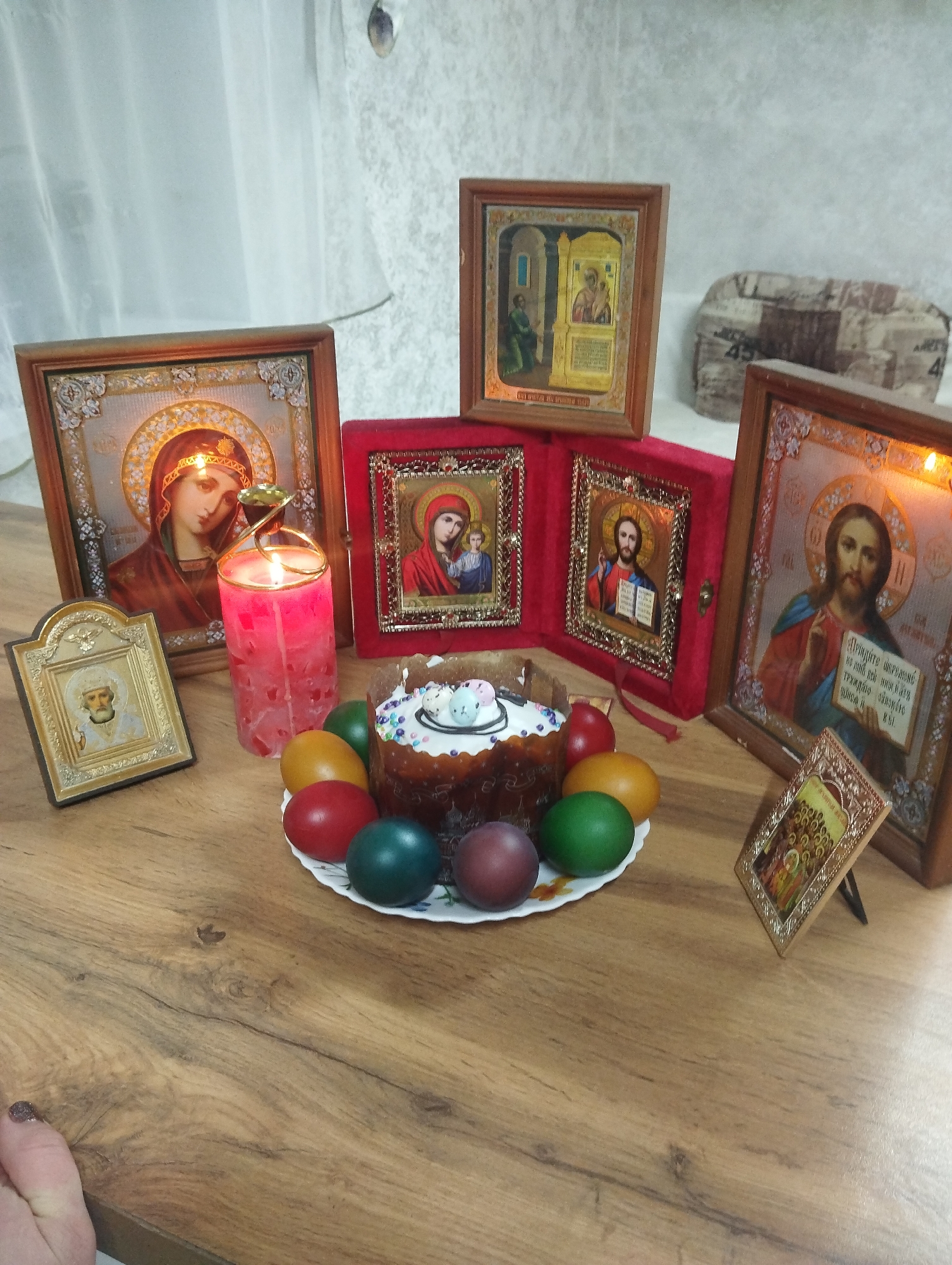 A wooden table with several religious icons, a candle, a plate of colored eggs, and a decorated Easter cake is visible in this image. A lit pink candle stands next to an icon of the Virgin Mary. Other icons are placed around the table, including one of Jesus Christ. The colored eggs are on a white plate surrounding the Easter cake, which is topped with white icing and small decorative eggs. The image was taken indoors, possibly in a home. The location may be near Grishanki, Russia.