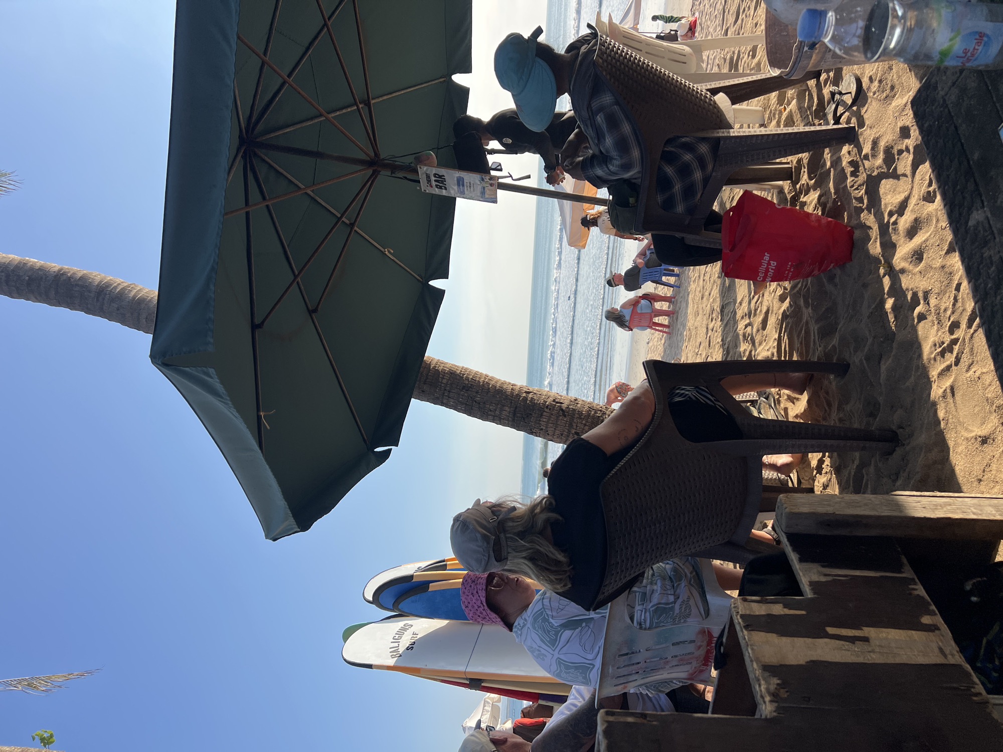 The image depicts a beach scene in Kuta, Indonesia, likely during daylight hours given the strong sunlight and clear blue sky. In the foreground, several people are seated at tables on the sand, some under a large green umbrella that provides shade. The individuals appear to be casually dressed, with some wearing hats. Surfboards are visible standing upright behind some of the seated patrons, suggesting proximity to surf lessons or rentals.
In the mid-ground, the ocean stretches out with gentle waves breaking on the shore. Further in the background, other beachgoers can be seen in the distance, some walking or sitting on the sand. A palm tree trunk is visible to the right of the frame, indicating a tropical environment.
The overall atmosphere is relaxed, consistent with a beachside establishment where people can enjoy the view and potentially refreshments, as hinted by a "BAR" sign hanging from the umbrella. The sand is coarse, typical of a beach, with footprints and shadows cast by the umbrella and furniture. A red reusable shopping bag with "CELLULAR WORLD" printed on it rests on the sand near one of the chairs. A partially visible bottle of "Le Mineral" water is in the lower right corner of the image.