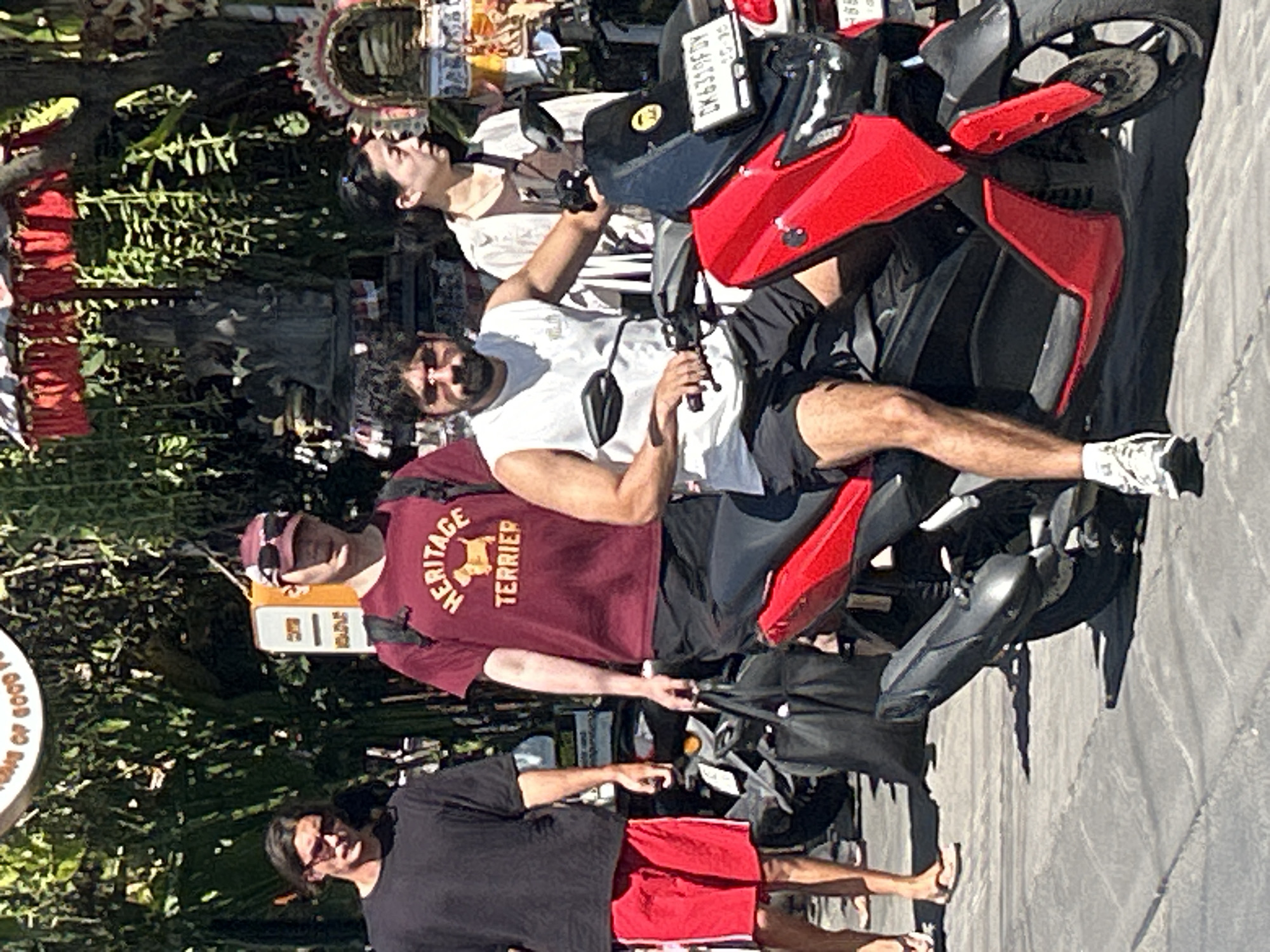 In North Kuta, Indonesia, four individuals are visible on a sunlit street. In the foreground, a man with dark hair and a beard is seated on a red and black scooter, wearing a white tank top and black shorts. His right hand is on the scooter's handlebar. To his left, a man with long dark hair and sunglasses is walking, dressed in a black t-shirt and red shorts. Behind the man on the scooter, a woman with dark hair pulled back is looking to her right, wearing a light-colored jacket over a brown shirt. To the left of the woman, another man with a dark baseball cap and sunglasses is walking, wearing a maroon t-shirt with the text "HERITAGE TERRIER" and black shorts. He is carrying two black bags in his left hand. Several parked scooters are visible in the background, along with lush green foliage and some decorative elements, suggesting a vibrant, possibly tourist-oriented area. The ground is paved with stone tiles.