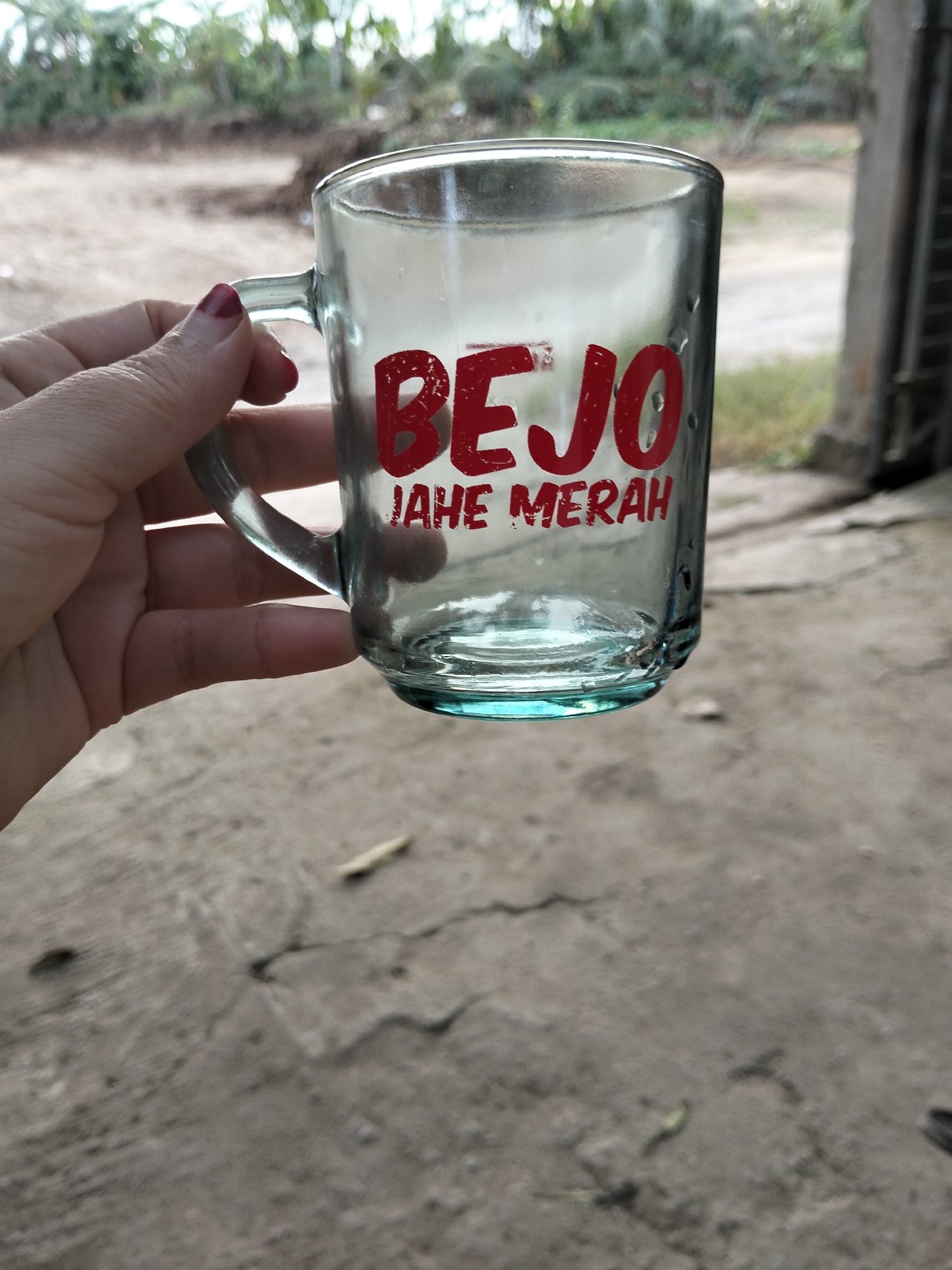 A close-up, eye-level, vertical shot shows a person's left hand with a painted fingernail holding a clear glass mug. The mug has red lettering that reads "BEJO JAHE MERAH". The background is blurred, showing greenery and a dirt path, consistent with an outdoor environment. The scene appears to be in a rural setting, possibly a yard or garden, in Payagaboh, Indonesia. The mug is the primary subject, positioned slightly off-center to the right. The overall lighting is natural daylight.