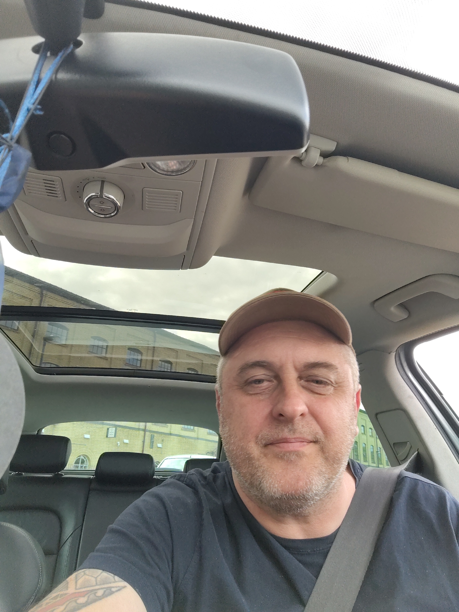 A middle-aged male individual with a light-colored beard and wearing a light brown baseball cap and a dark t-shirt is seated in the driver's seat of a vehicle, looking directly at the camera. He is wearing a grey seatbelt, and a multi-colored tattoo is visible on his left forearm. The vehicle's interior features black leather rear seats and a grey overhead console with controls for functions like a panoramic sunroof. A rearview mirror with blue strings or ribbons attached hangs in the upper left. Through the panoramic sunroof and rear window, a large, light-colored brick building with multiple evenly spaced windows, some with arched tops, is prominently visible under an overcast sky. This architectural style is consistent with the redeveloped industrial buildings located in the Fletton Quays area of Peterborough, United Kingdom. A white vehicle is partially visible in the distance behind the main building.