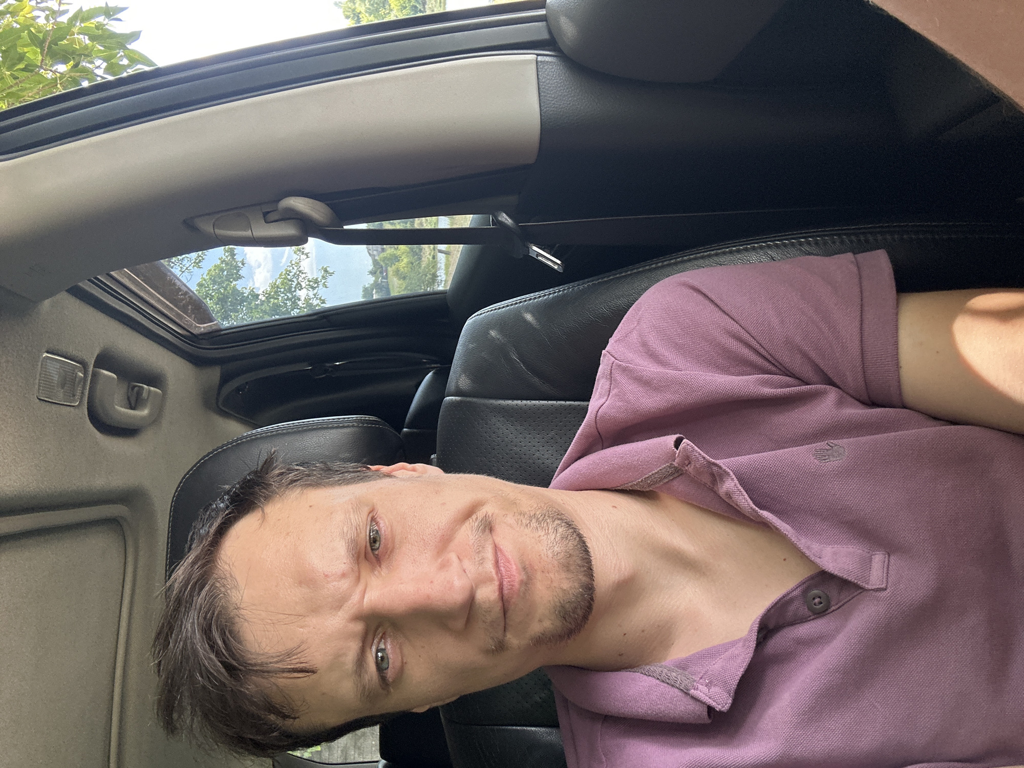 A medium shot selfie shows a man sitting in the driver's seat of a vehicle, with his face filling the majority of the frame. He has short, brown, messy hair and a light beard. His eyebrows are furrowed, and he is looking directly at the camera with a neutral expression. He is wearing a purple polo shirt with a collar and a button at the neck. The interior of the car is visible, including the black leather seat, the car's roof, and the passenger side door. Outside the car, through the window, a body of water and trees can be seen under a bright sky. The location is identified as Makiv, Ukraine.