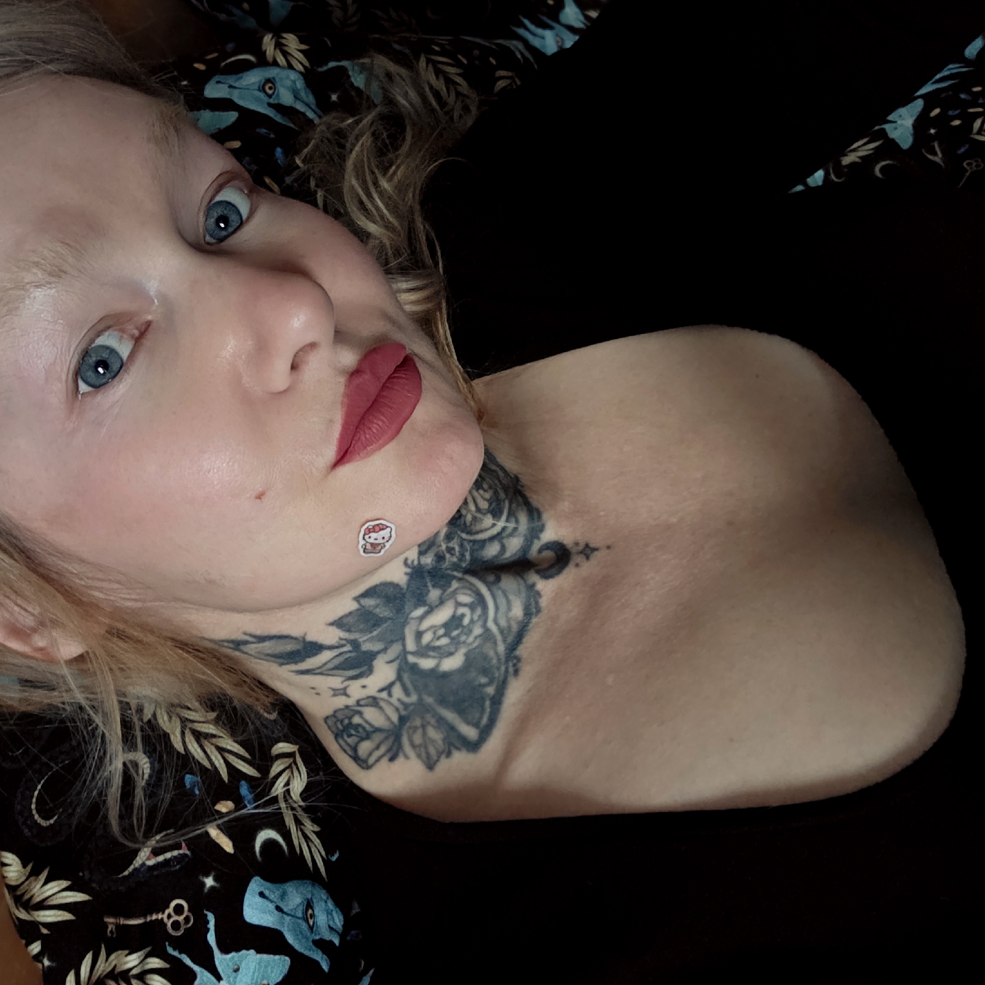 A selfie of a light-skinned person with blue eyes and blonde hair lying down. They have a floral tattoo on their neck and a "Hello Kitty" sticker on their cheek. They are wearing red lipstick and are positioned against a black background with a dark patterned fabric visible on one side. The environment cannot be confirmed as Saint Petersburg, Russia.