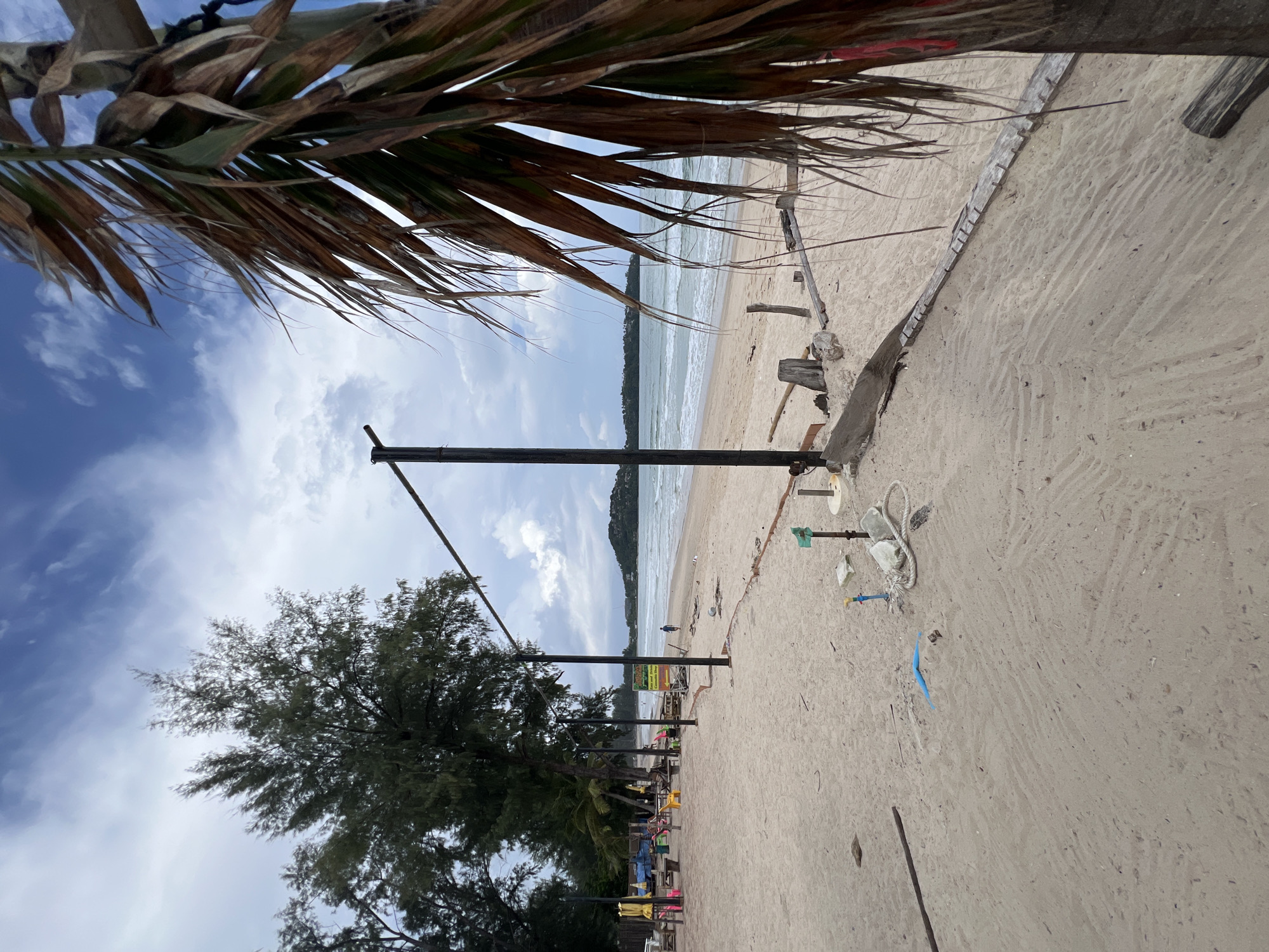 The image depicts a beach scene in Choeng Thale, Thailand, under a cloudy sky with patches of blue. The foreground is dominated by a sandy beach with a visible volleyball net pole and a large palm tree frond to the right. Scattered on the sand are pieces of driftwood, some blue plastic tubing, a white rope, a metal post with a green flag, and a blue spigot-like object. In the mid-ground, there is a line of trees, some beach chairs and umbrellas, and a sign. Further in the background, the ocean waves are visible, and a distant island or hill is seen on the horizon. A single person is walking on the beach in the distance. The overall impression is that of a somewhat weathered or less-than-pristine beach environment, possibly indicating it is off-season or undergoing maintenance.