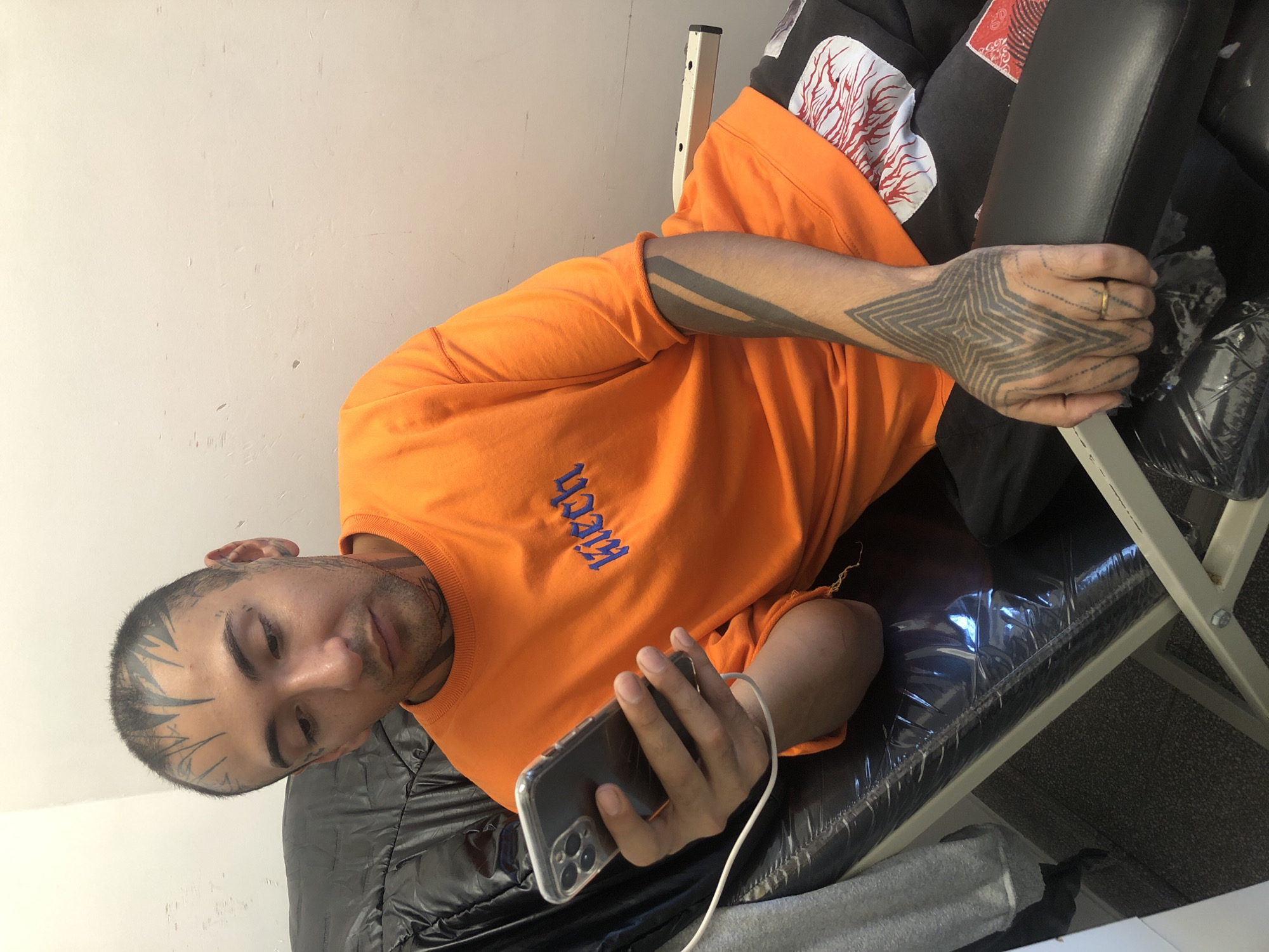 A medium shot depicts a light-skinned man with visible tattoos on his head, arms, and hands. He is wearing an orange t-shirt with the word "Hitch" embroidered in blue letters. He is lying on a black, padded treatment table or chair, and he is holding a smartphone connected to a white charging cable. The background consists of a plain white wall on the left side of the image and a glimpse of the surrounding objects on the right, including what appears to be furniture. The setting seems to be indoors, possibly a tattoo studio or a similar type of treatment room. There is no evidence that the photograph was taken in Buenos Aires, Argentina.