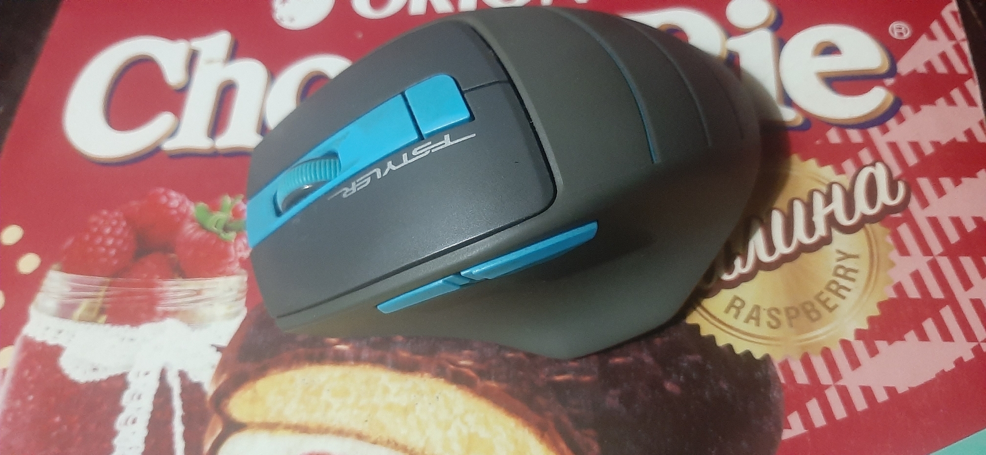 The image shows a gray and blue computer mouse placed on a red surface with a pattern. The surface appears to be a food packaging, as evidenced by the text "Raspberry" and what looks like images of berries and cake. The mouse has a scroll wheel and two additional blue buttons on the side. The text "FSTYLER" is visible on the mouse. The lighting is dim, casting shadows. The image is a close-up shot, with the mouse occupying the majority of the frame. There is no indication of the location being Yessentukskaya, Russia, as the scene is indoors and does not contain any recognizable landmarks or context clues that would suggest this specific location.