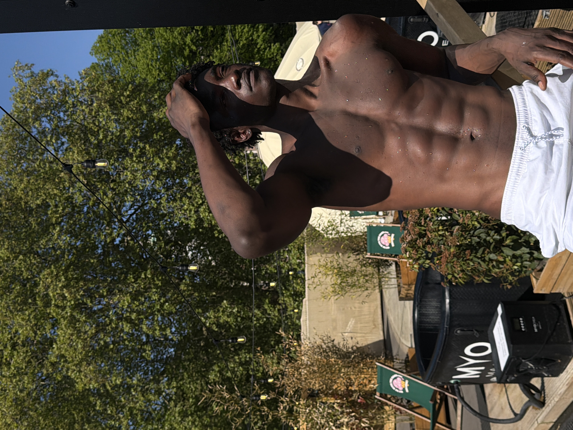 The image shows a shirtless man with dark skin, wet from what appears to be sweat or water, standing outdoors on what appears to be a wooden deck. He is wearing white shorts and has one hand on his head. To the side is a black tub labeled "MYO" and a black machine connected to it. In the background, there are green trees, string lights, and green chairs with a logo. The location appears to be an outdoor event space in London, United Kingdom.