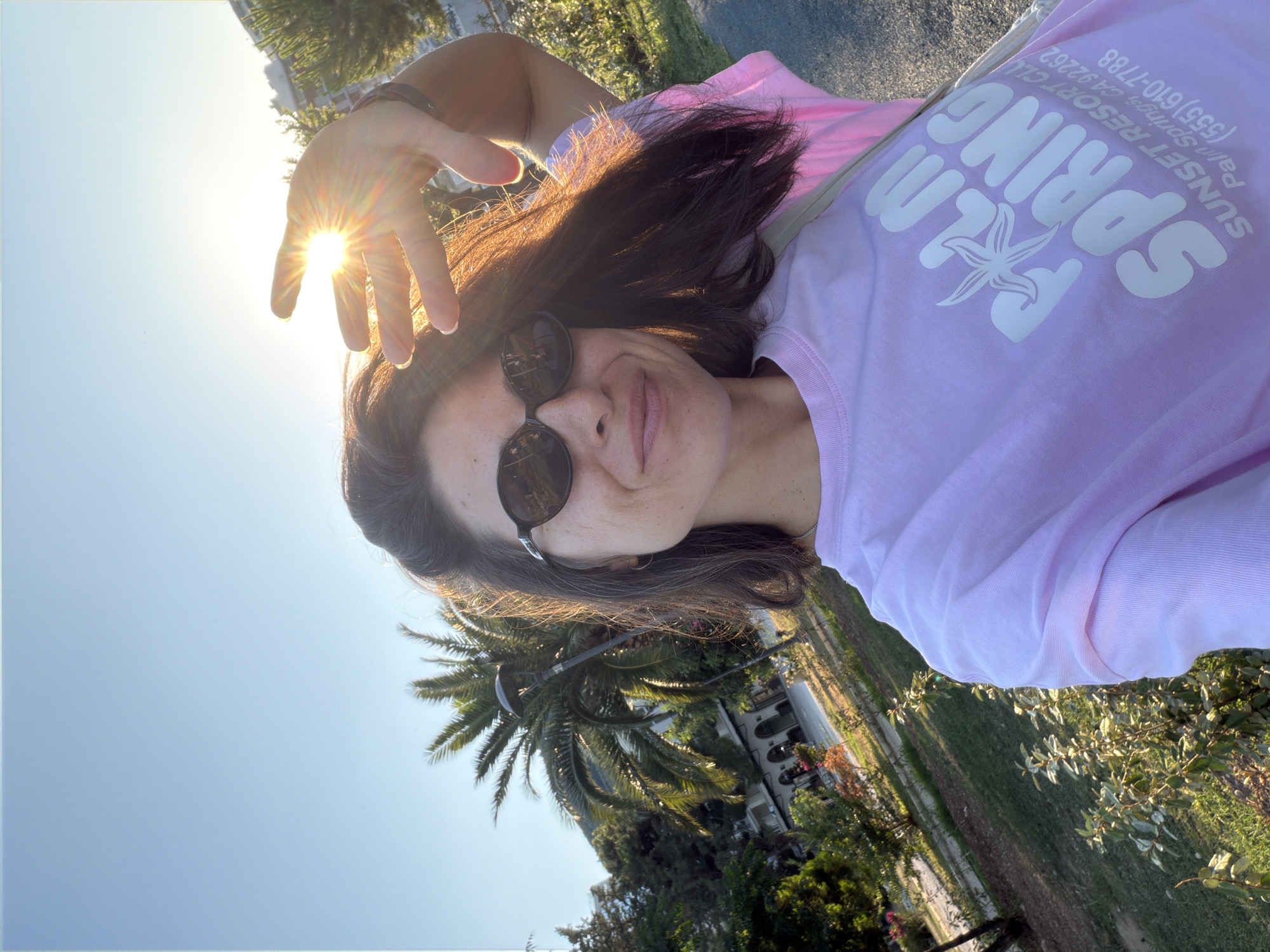 A woman is taking a selfie outdoors, with the sun shining brightly. She is wearing a light purple t-shirt with "PALM SPRING" printed in white lettering, along with a starfish graphic. The t-shirt also has text that appears to be "SUNSET RESORT CLUB Palm Springs, CA 92262 (555) 610-7788". She has on dark sunglasses and her dark hair is down. Her left hand is raised, partially obscuring the sun. The background shows lush green foliage, including palm trees, and what appears to be a building. The bright sunlight suggests it is daytime, likely midday or afternoon. The presence of palm trees and the mention of "Palm Springs" on her shirt hint at a warm, possibly tropical or Mediterranean climate, consistent with the provided location context of Budva, Montenegro. The woman's expression seems relaxed and cheerful, enjoying the sunny weather.