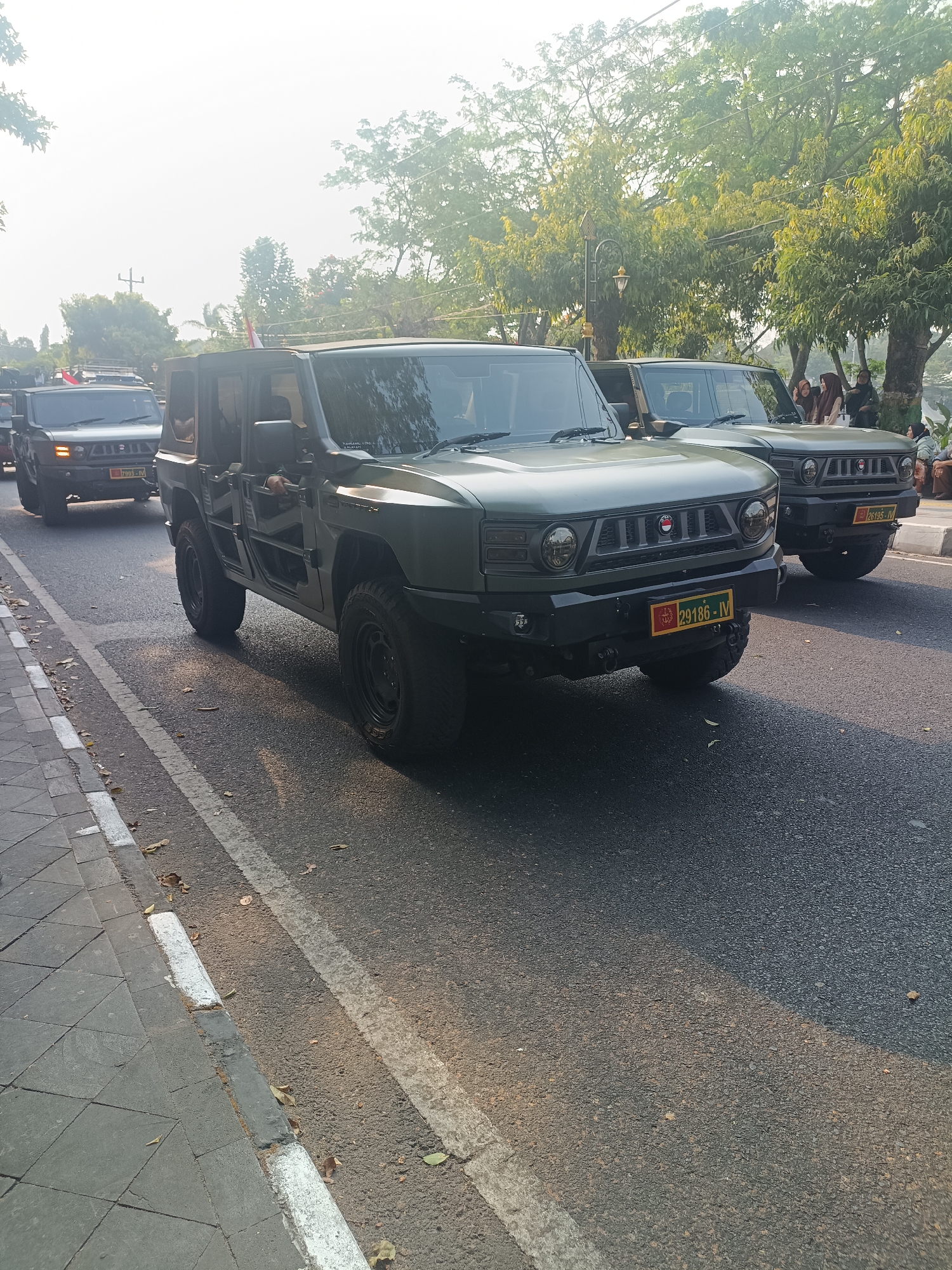 In Sleman, Indonesia, two olive green military-style SUVs are driving on an asphalt road. The lead vehicle, on the right, has its doors removed and is driven by a person wearing a dark shirt. The second vehicle, on the left, is closer to the camera and appears to be a similar model. Both vehicles have dark colored wheels and matte black bumpers. The lead vehicle displays a yellow license plate with black text and insignia. The second vehicle, partially visible behind the first, also has a yellow license plate with black lettering. The road has white lane markings. To the left of the road, there is a tiled sidewalk with a white-bordered curb. In the background, trees with green foliage are visible, and on the right side of the frame, several people, including women wearing headscarves, are standing near the trees. The sky is bright and hazy, suggesting it is daytime.