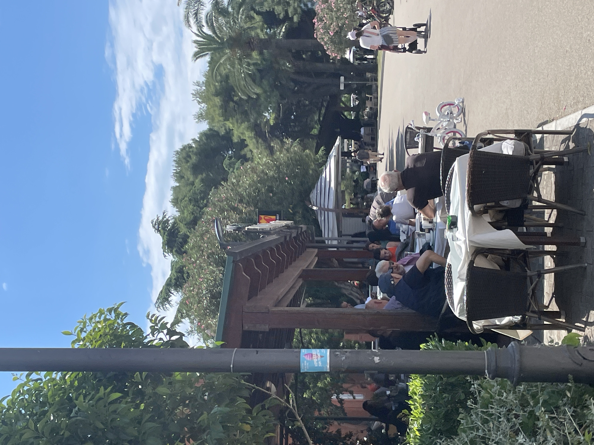 The image shows an outdoor dining area at a cafe, likely in Bar, Montenegro. Several people are seated at tables with white tablecloths, some are visible eating or conversing. The cafe has a wooden pergola-style structure. A street runs alongside the dining area with pedestrians walking and cycling. Lush green vegetation is abundant. The sky is bright blue with scattered white clouds.