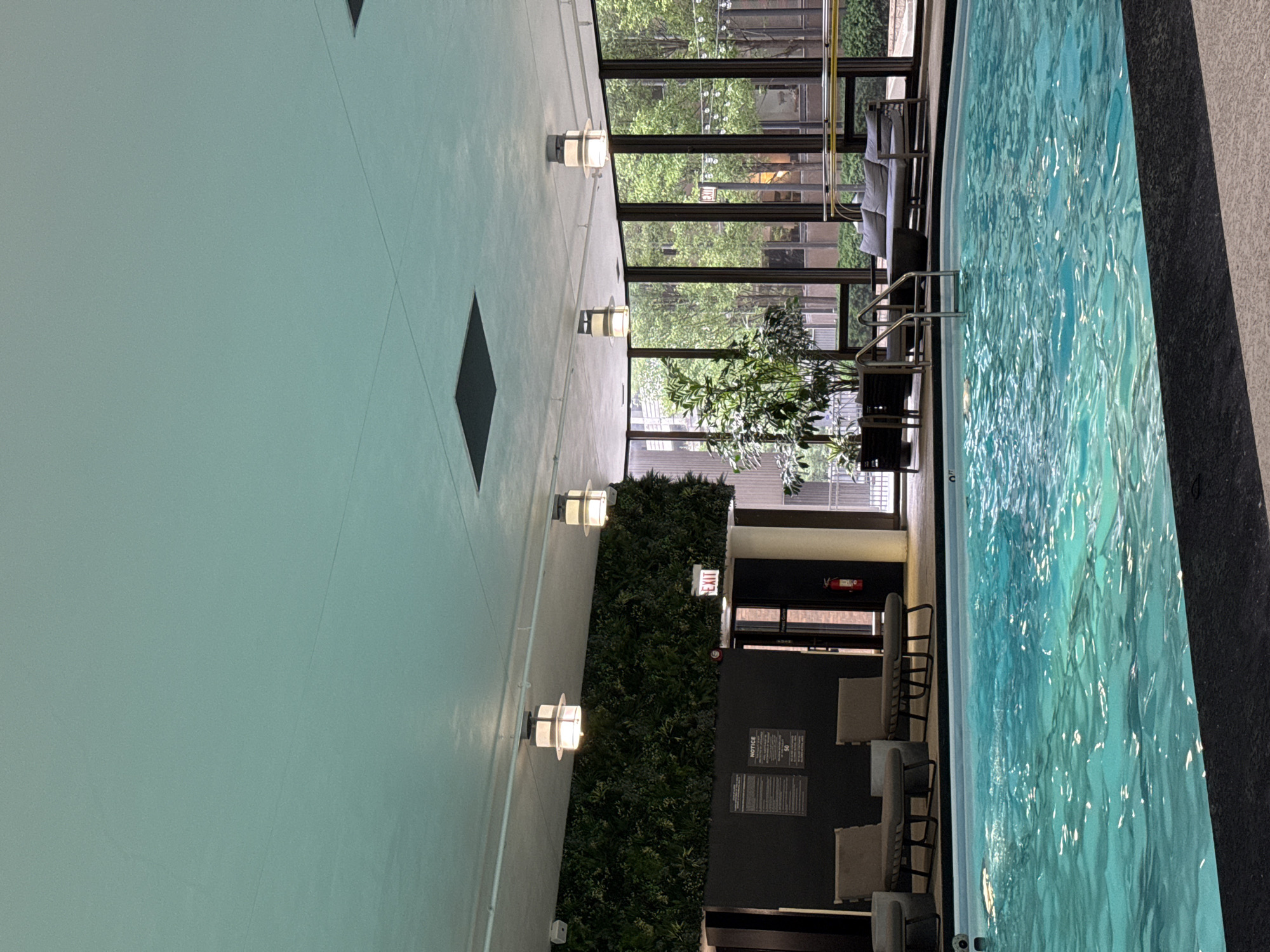 This image depicts an indoor swimming pool area located in Chicago, Illinois, United States of America. The pool is illuminated by overhead lighting and the sunlight filtering through large windows that offer a view of trees and buildings outside. The water in the pool is a vibrant turquoise color and appears to be calm, with ripples visible on the surface.
Along the edge of the pool, there are lounge chairs and a small table, suggesting a relaxation area. To the left of the pool, a wall is adorned with a large artificial green plant installation, creating a naturalistic aesthetic. Above this green wall, modern ceiling lights are visible.
Further to the left, there are signs posted on a dark wall, including a notice with text and an "EXIT" sign above a doorway. A fire extinguisher is also mounted on the wall near the exit. In the background, through the windows, one can observe the exterior environment, which includes more greenery and architectural elements of the city. A seating area with a couch and a coffee table is also visible near the windows. The ceiling is a light aqua color with some square-shaped recesses and visible lighting fixtures.