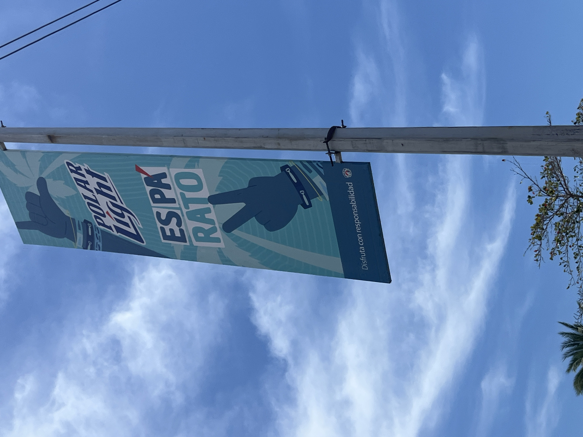 A horizontal banner hangs from a metal pole against a bright blue sky with wispy clouds. The banner advertises "Polar Light" and the phrase "ESPA RATO" in large, bold letters, accompanied by two stylized blue hands making hand gestures. A small logo is visible on the right side of the banner, along with the Spanish phrase "Disfruta con responsabilidad" (Enjoy with responsibility). The pole also has power lines at the top and part of a tree is visible on the right, including branches with foliage and the frond of a palm tree. The location is in the city of Unknown, Unknown.