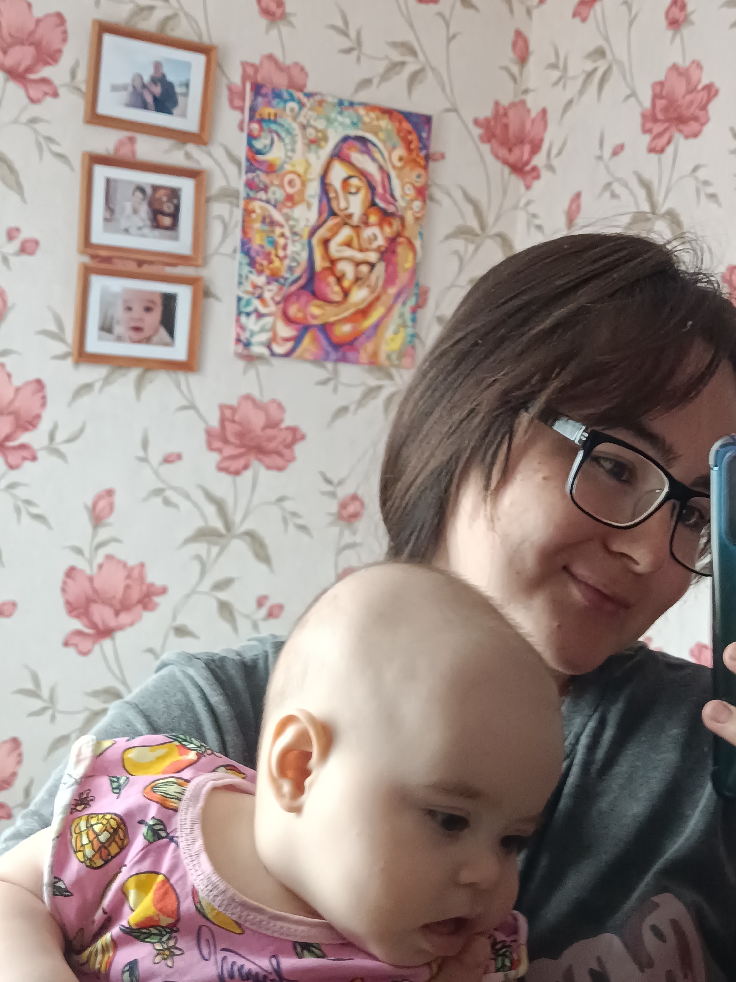 A woman wearing glasses and a gray shirt is holding a baby in a pink shirt with yellow fruit designs. The woman is holding up a phone and appears to be taking a selfie. The background features a floral wallpaper with large pink flowers. There are also three framed photos and a painting visible on the wall behind the woman. The frames are arranged in a vertical line. The specific location in Noviy, Russia cannot be determined from the image.